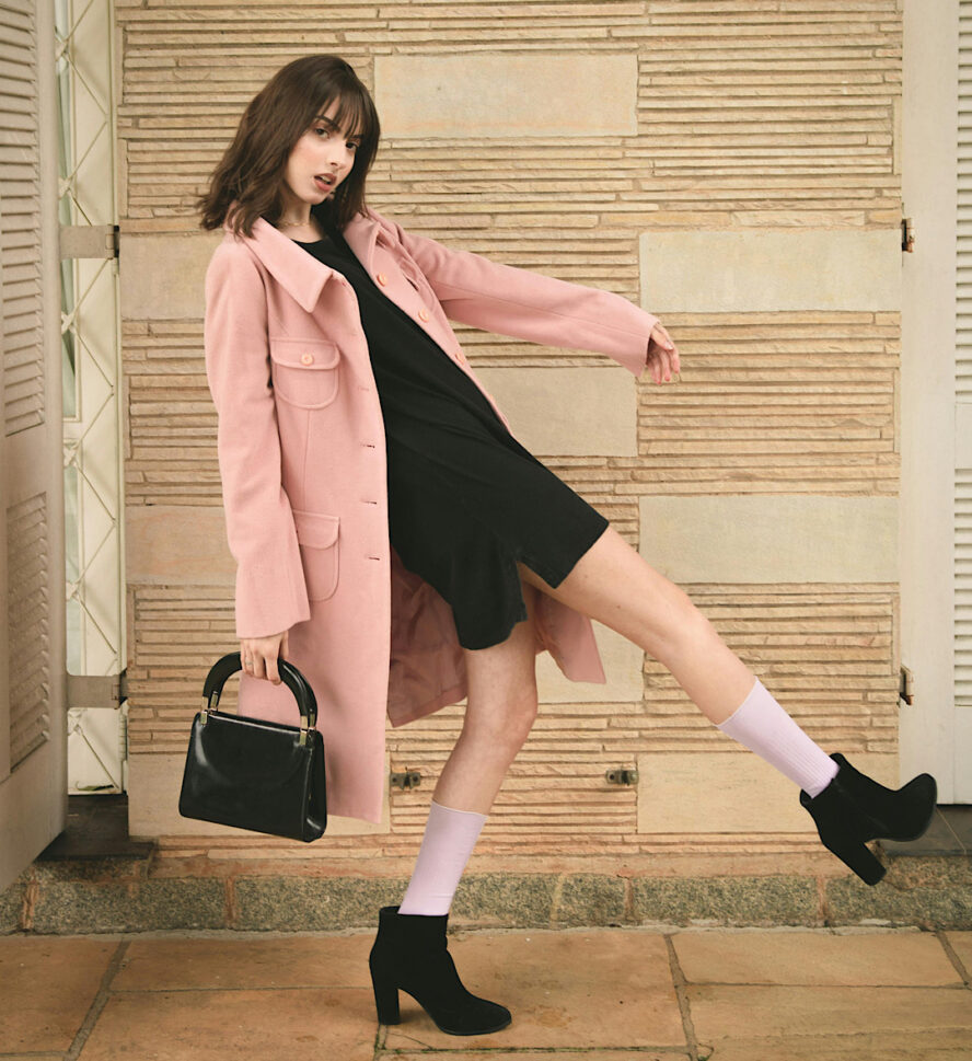 Young woman in a pink overcoat with a black dress, handbag, and shoes walking is an exaggerated way.