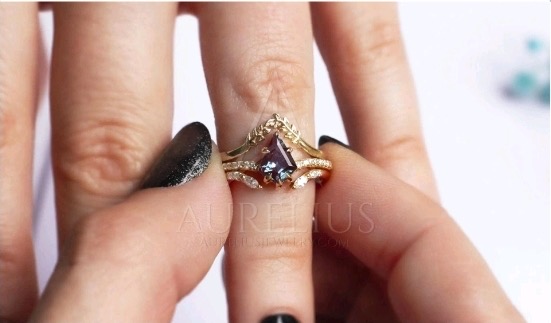 Close-up on a woman’s hands, where she holds a ring on her finger. The woman wears black nail polish, and the golden ring has crystals and a stone.