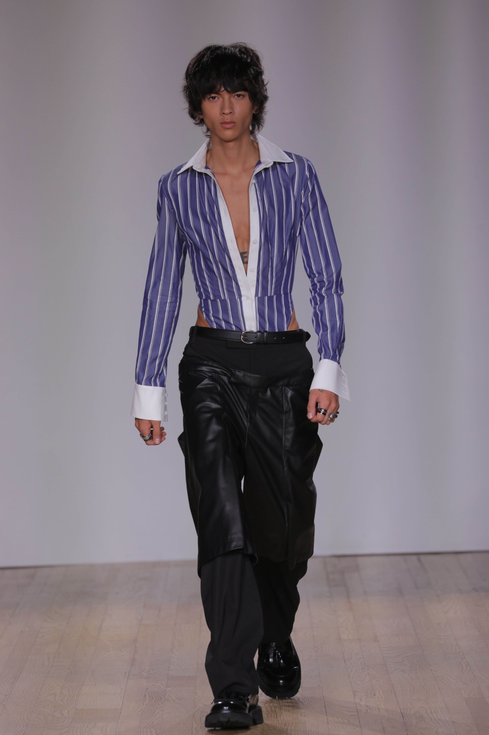 A model walking the runway a blue pinstripe shirt and black pants.