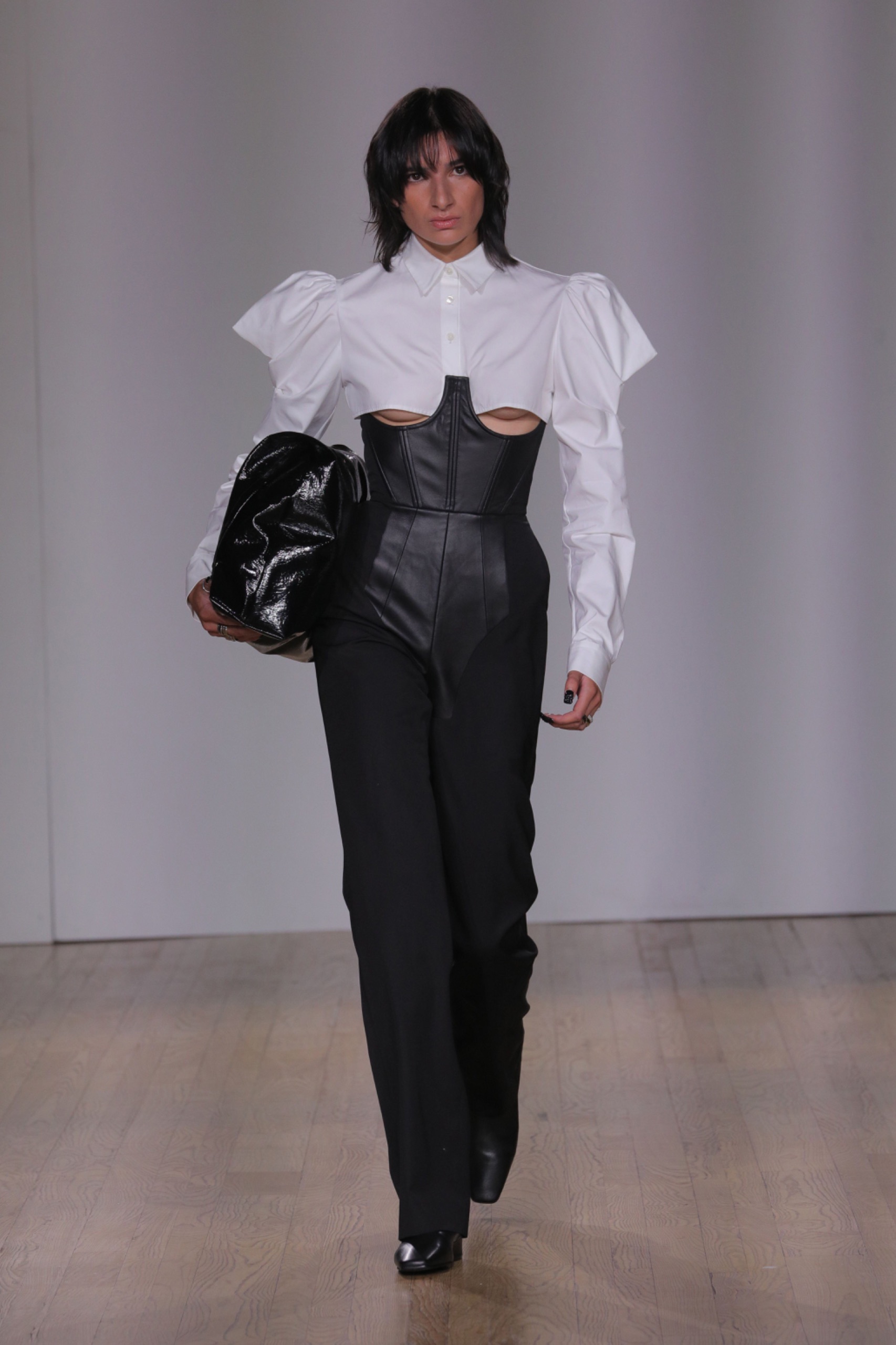 A model walking the runway wearing a cropped white shirt, a black corset, black bag and black bottoms.