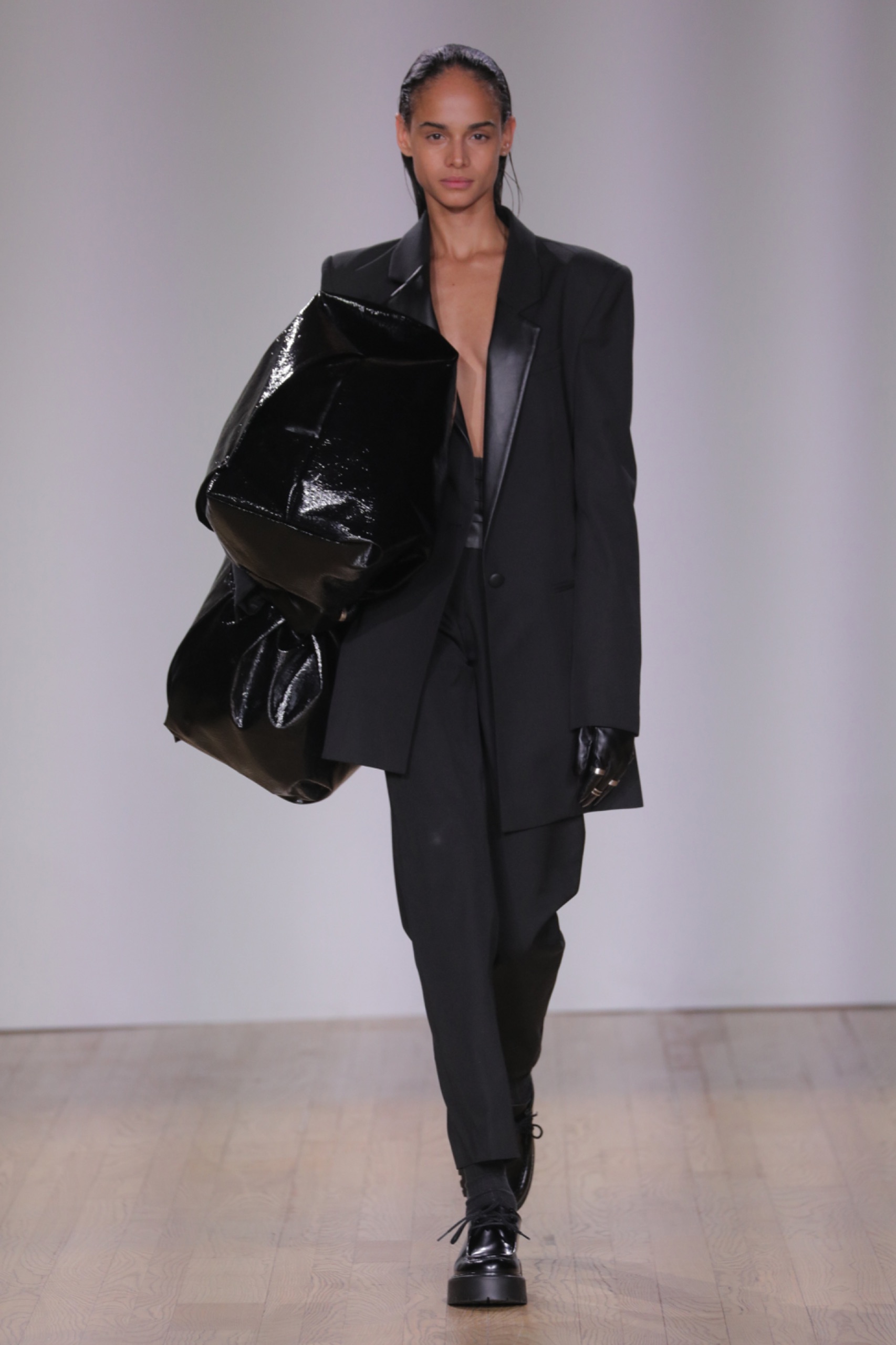 A model walking the runway wearing a black blazer, bag and bottoms.