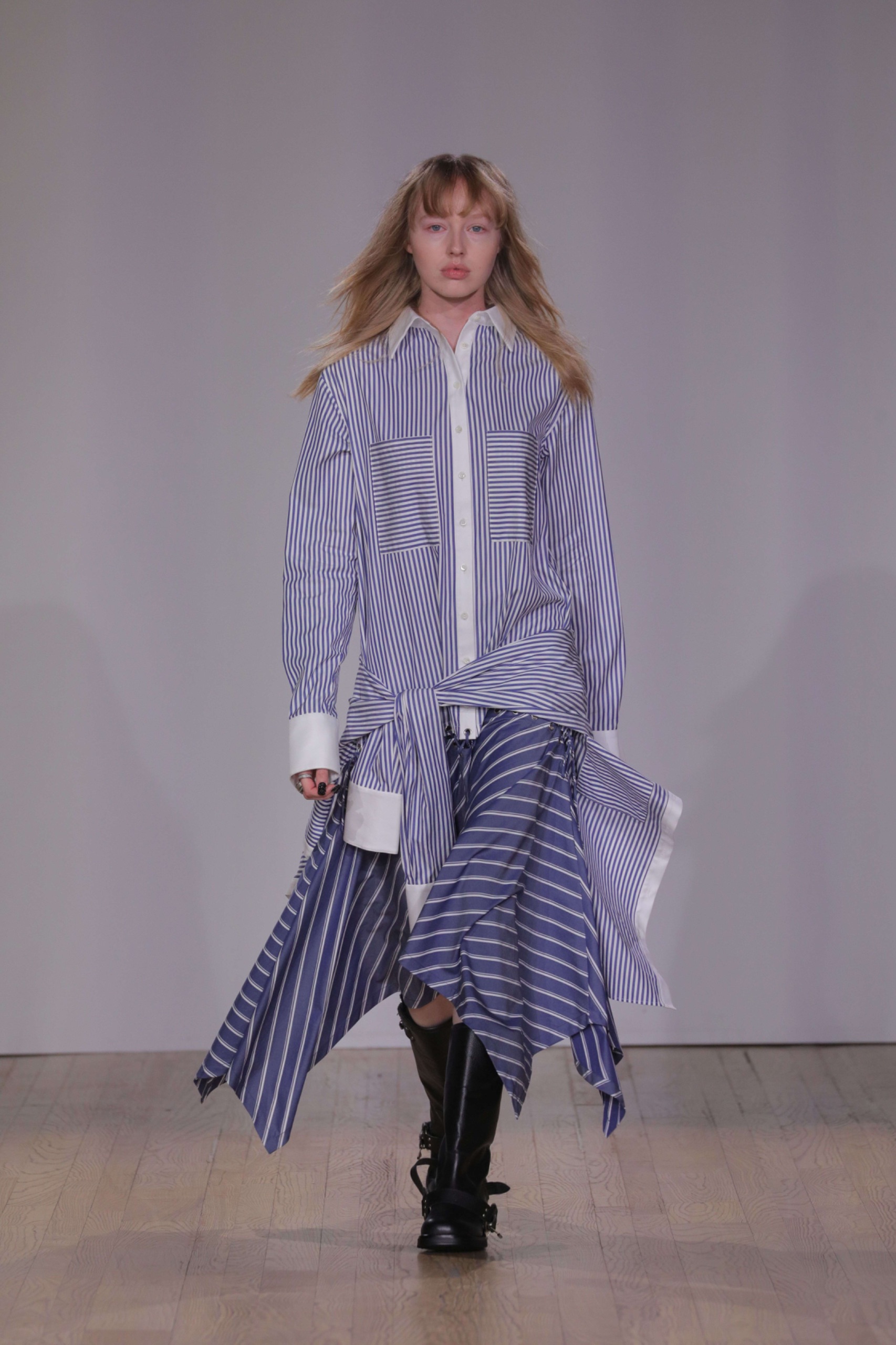 A model walking the runway wearing a long blue pinstripe top and matching skirt with black boots.