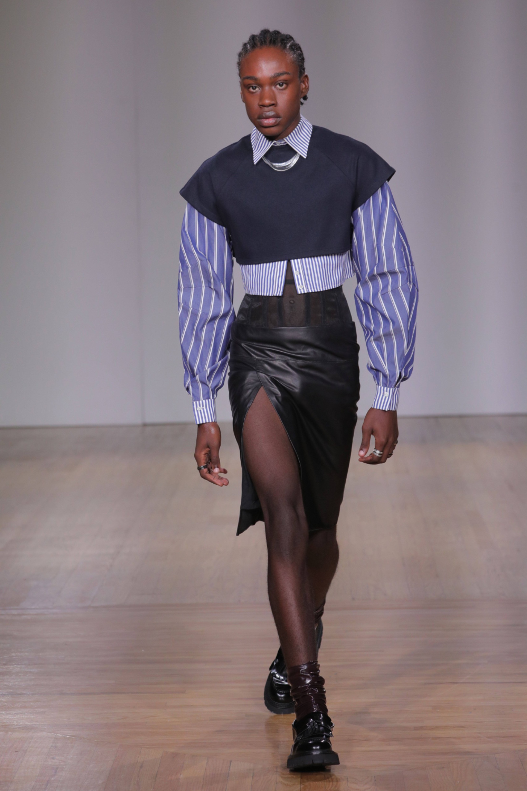 A model walking the runway wearing a long sleeved blue pinstripe shirt, a cropped black jumper and blue skirt.