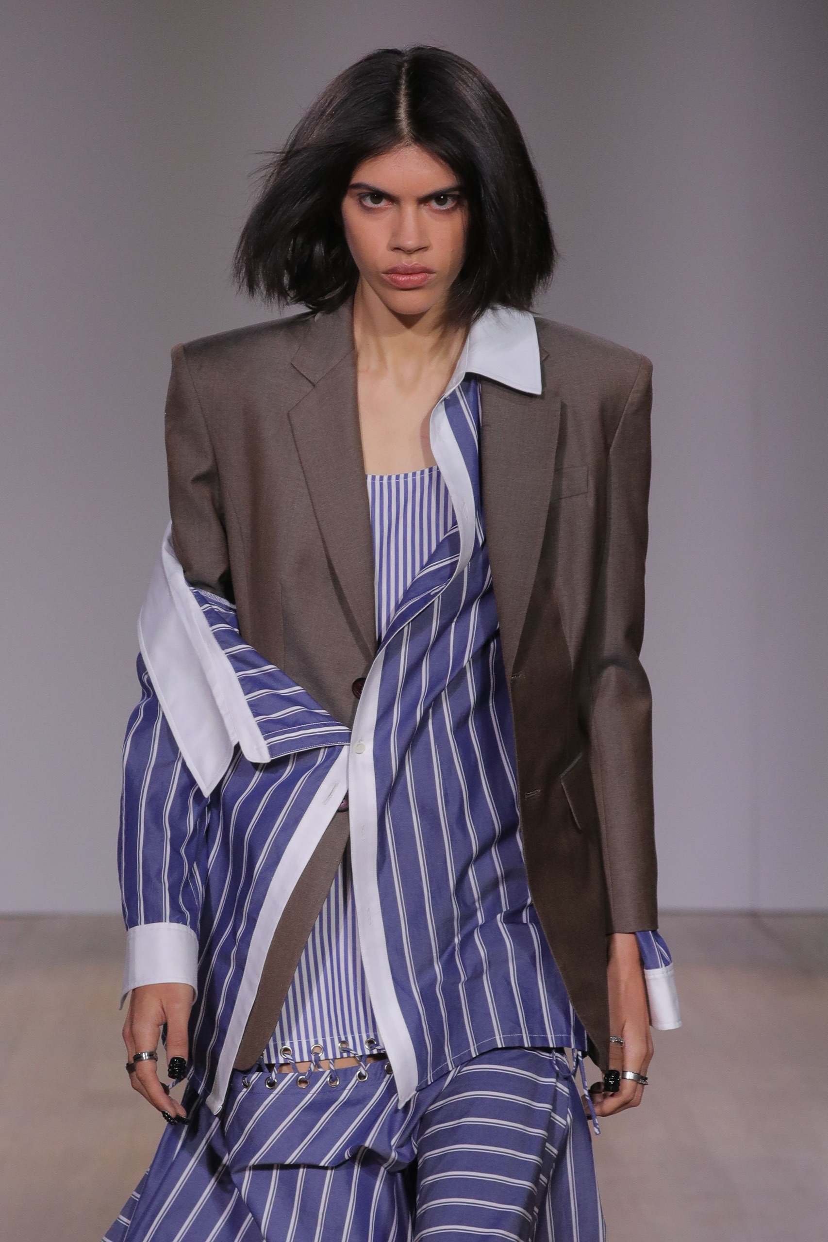 A model walking the runway wearing blazer and blue pinstripe dress.