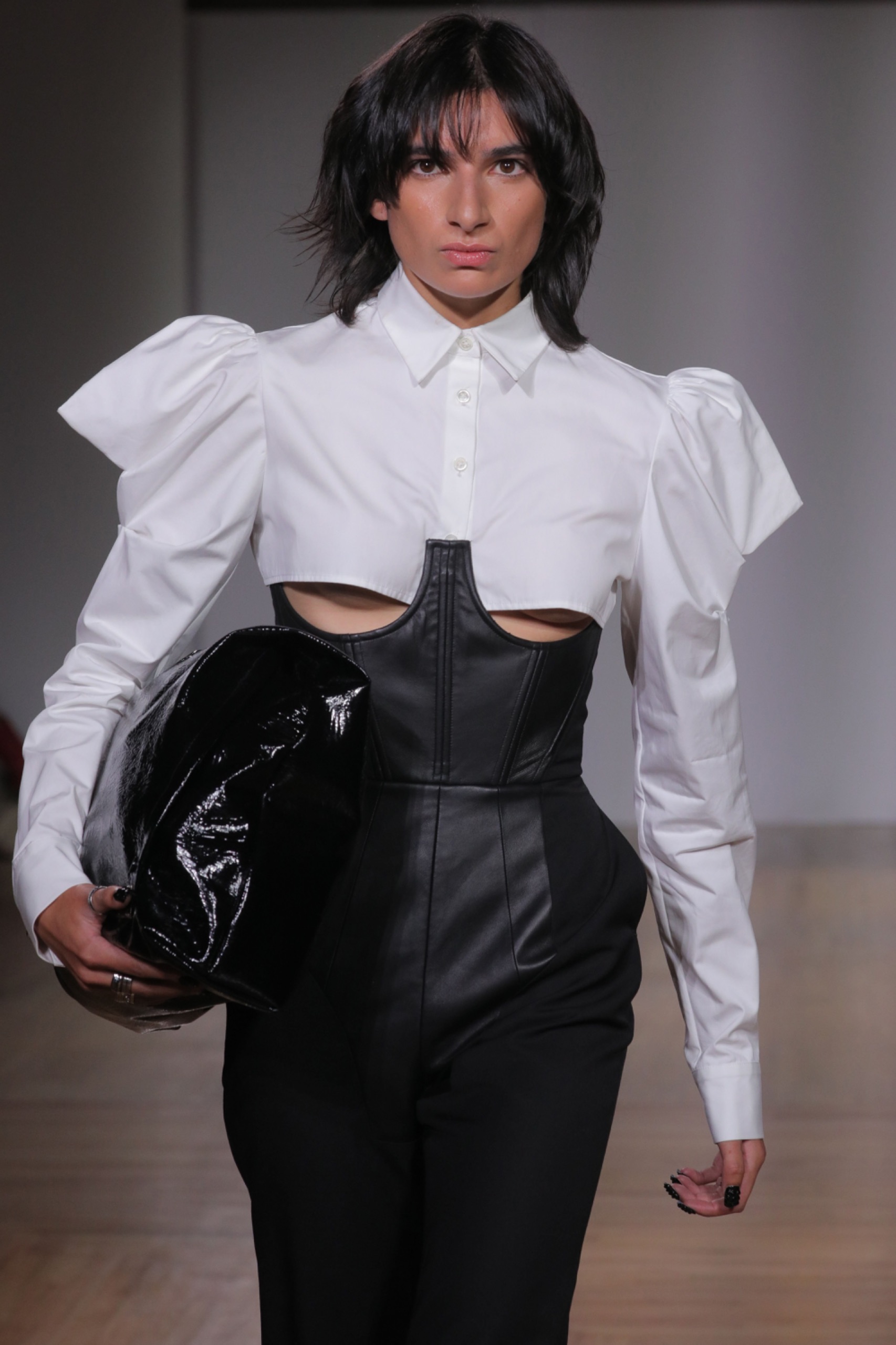 A model walking the runway wearing a cropped white shirt, a black corset, black bag and black bottoms.