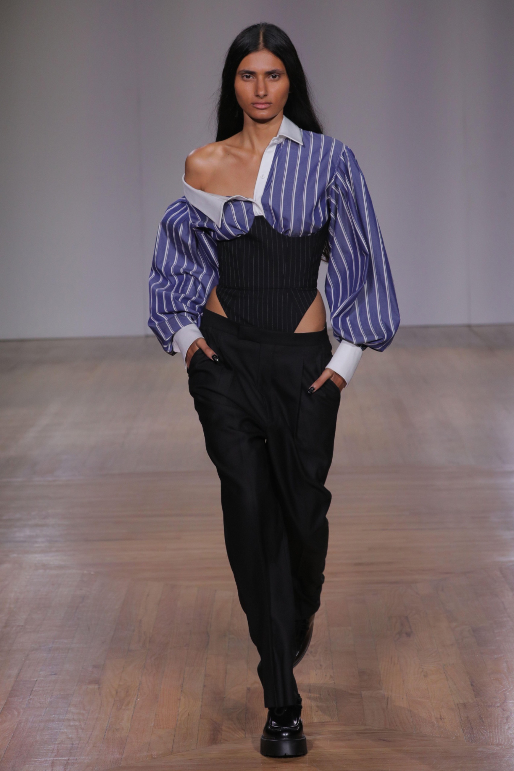 A model walking the runway wearing a blue pinstripe shirt, black corset and black bottoms.