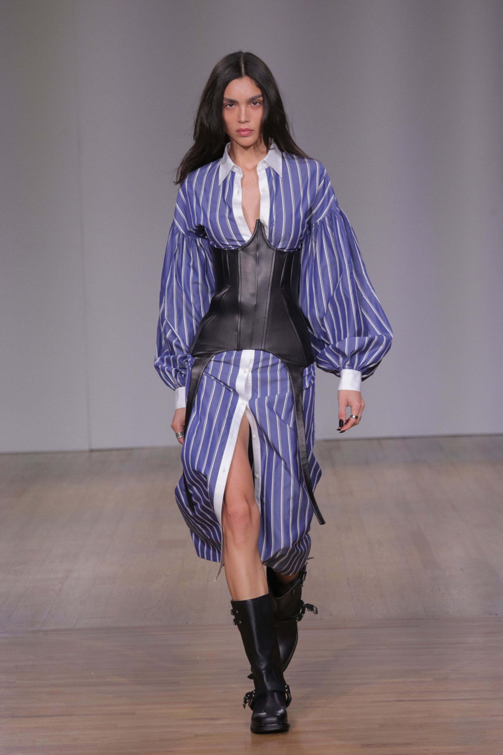 A model walking the runway wearing a blue pinstripe dress with black corset and black shoes.