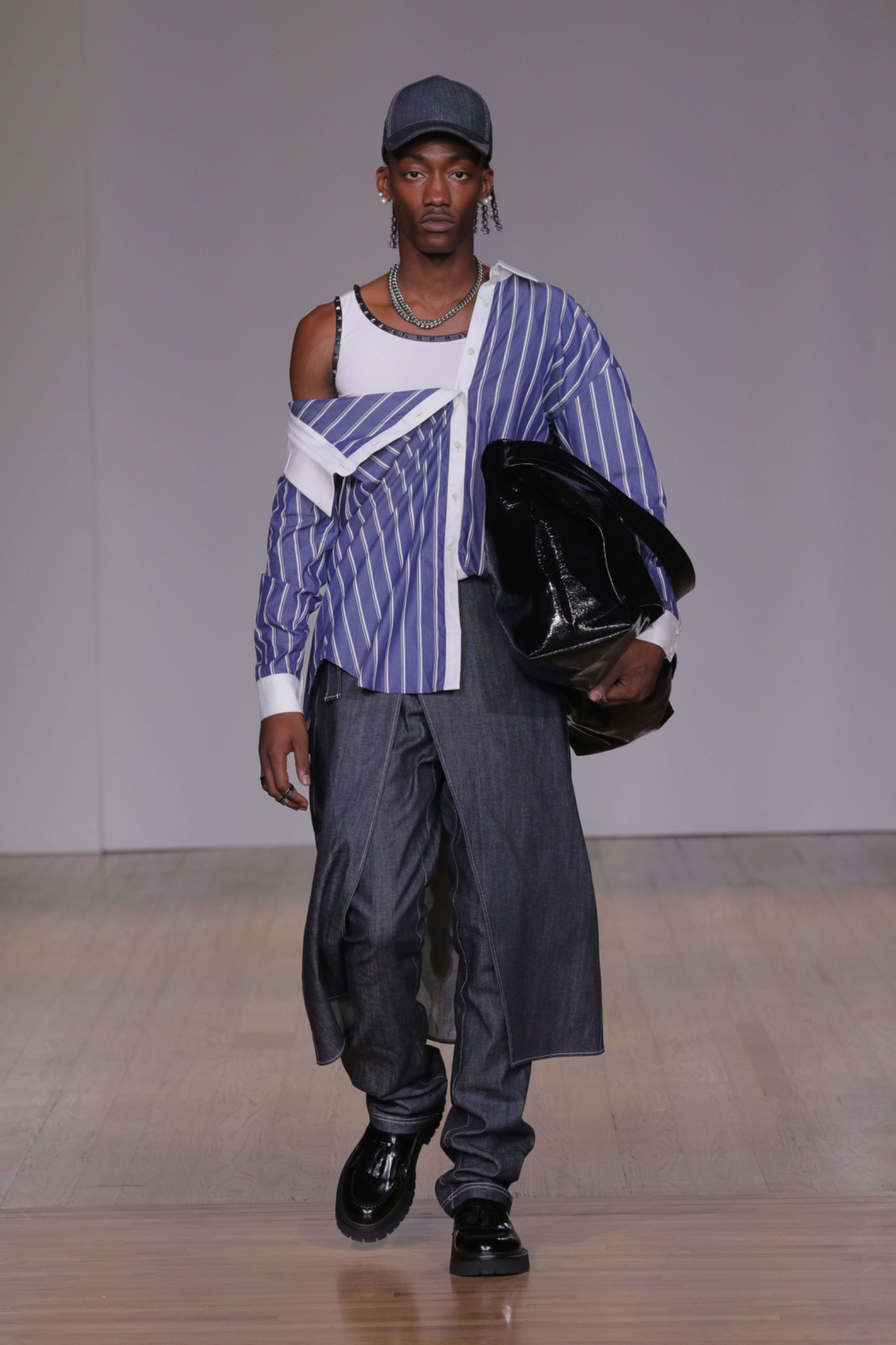 A model walking the runway wearing a white vest, blue pinstripe shirt, grey trousers and a black bag.