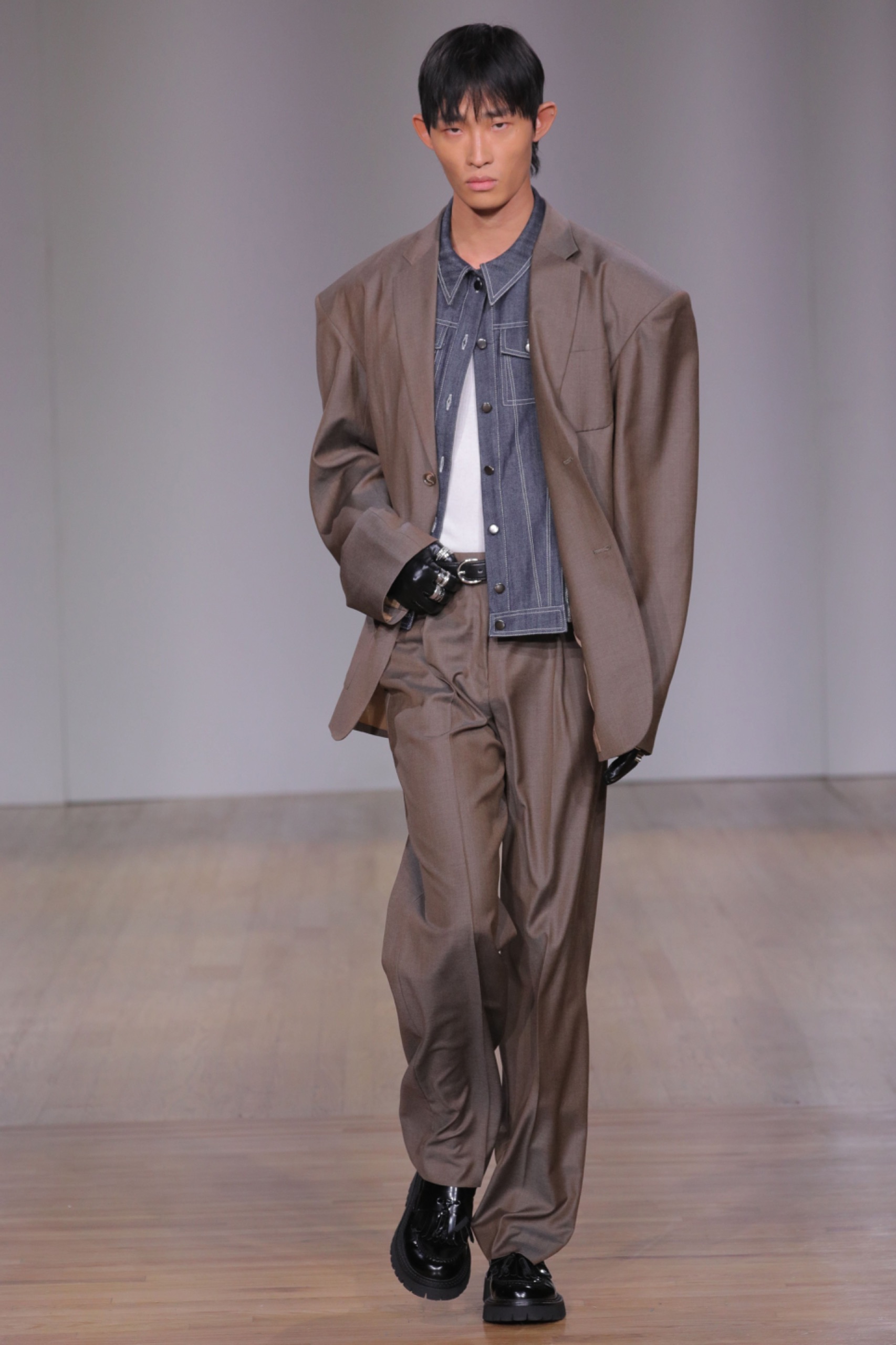 A model walking the runway wearing an oversized brown suit with denim shirt.