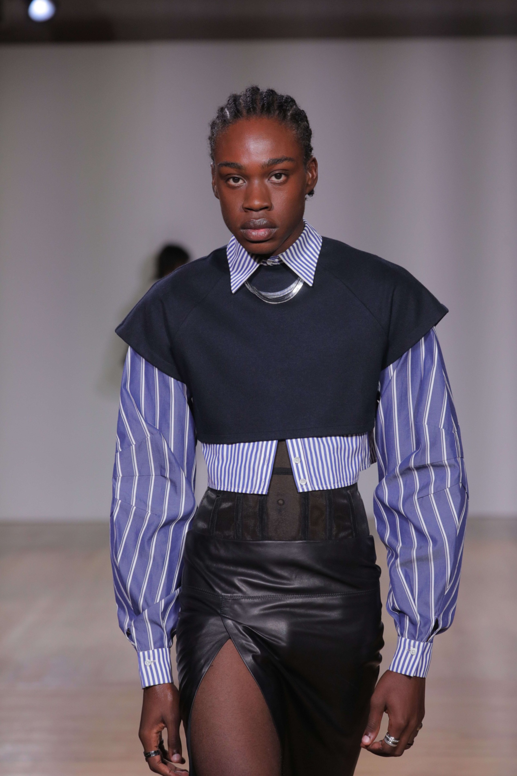 A model walking the runway wearing a black vest over a blue pinstriped shirt with a leather skirt.