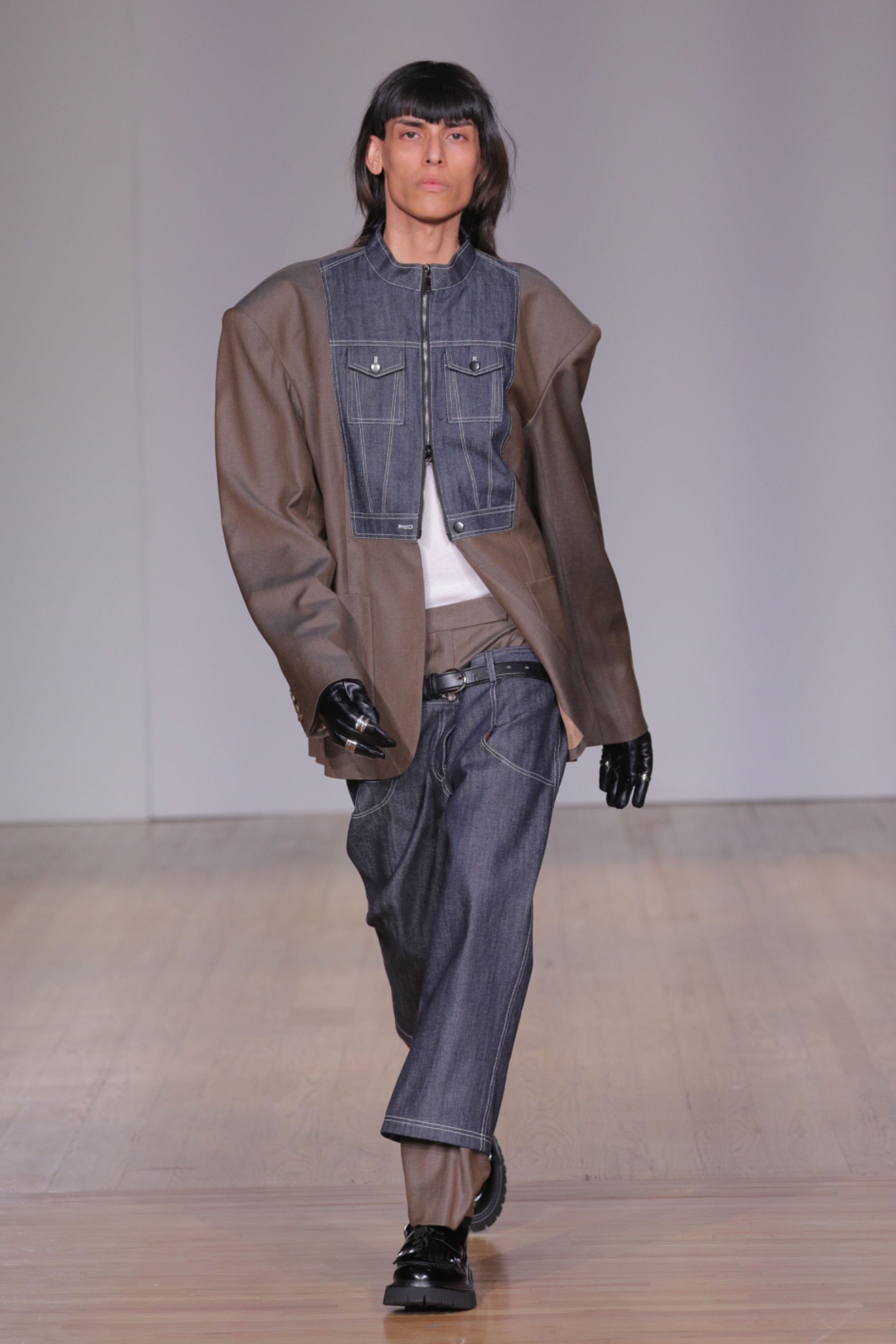 A model walking the runway wearing a brown jacket, with jeans and denim vest.