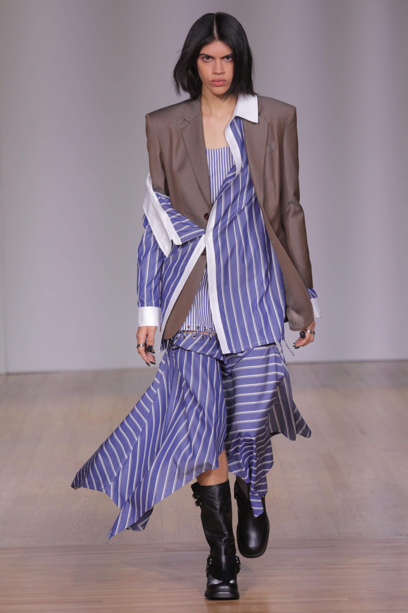 Model walking the runway wearing a light brown blazer with black shoes and blue and white striped skirt and matching shirt.