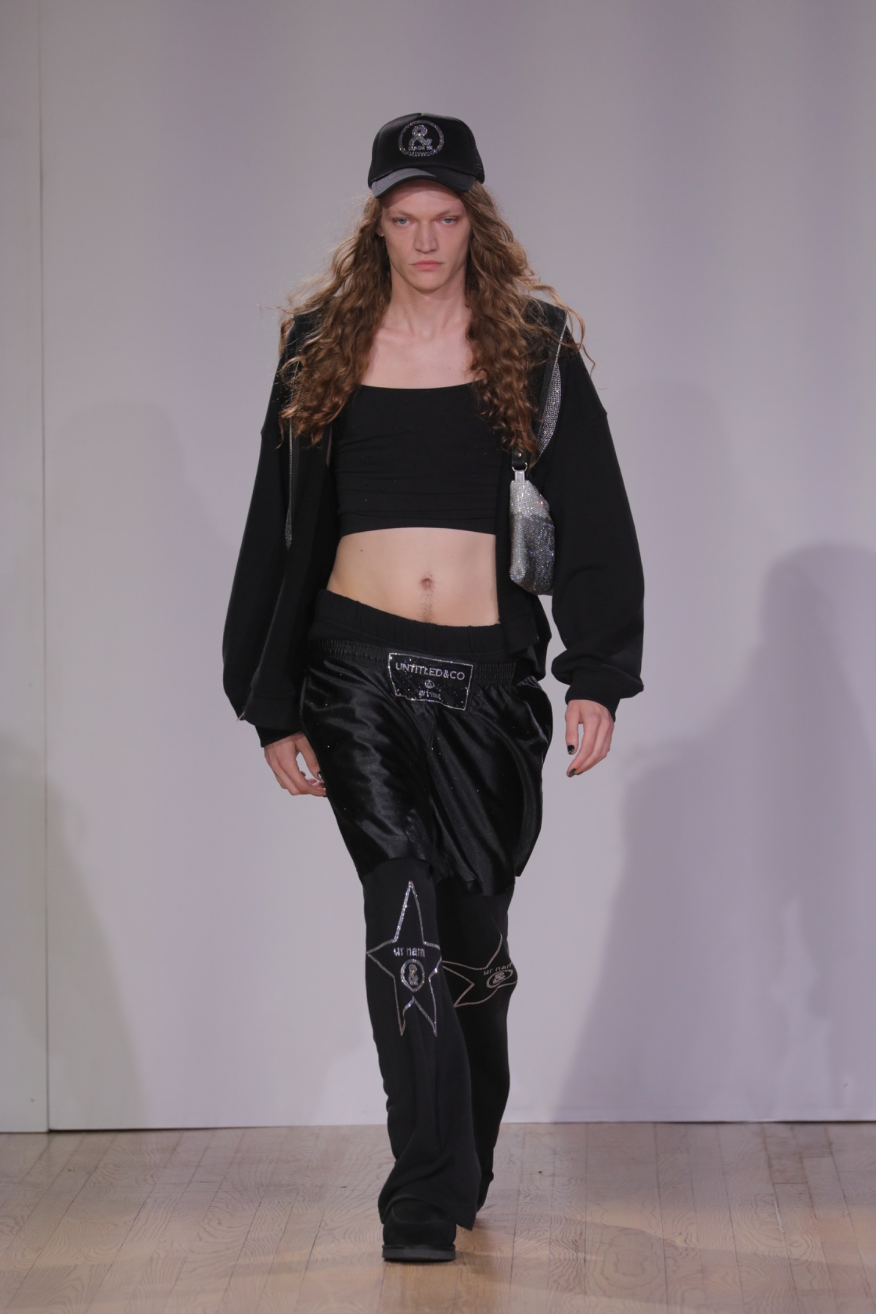 A model walking the runway wearing a black cap, black top, black pants and a black jacket.