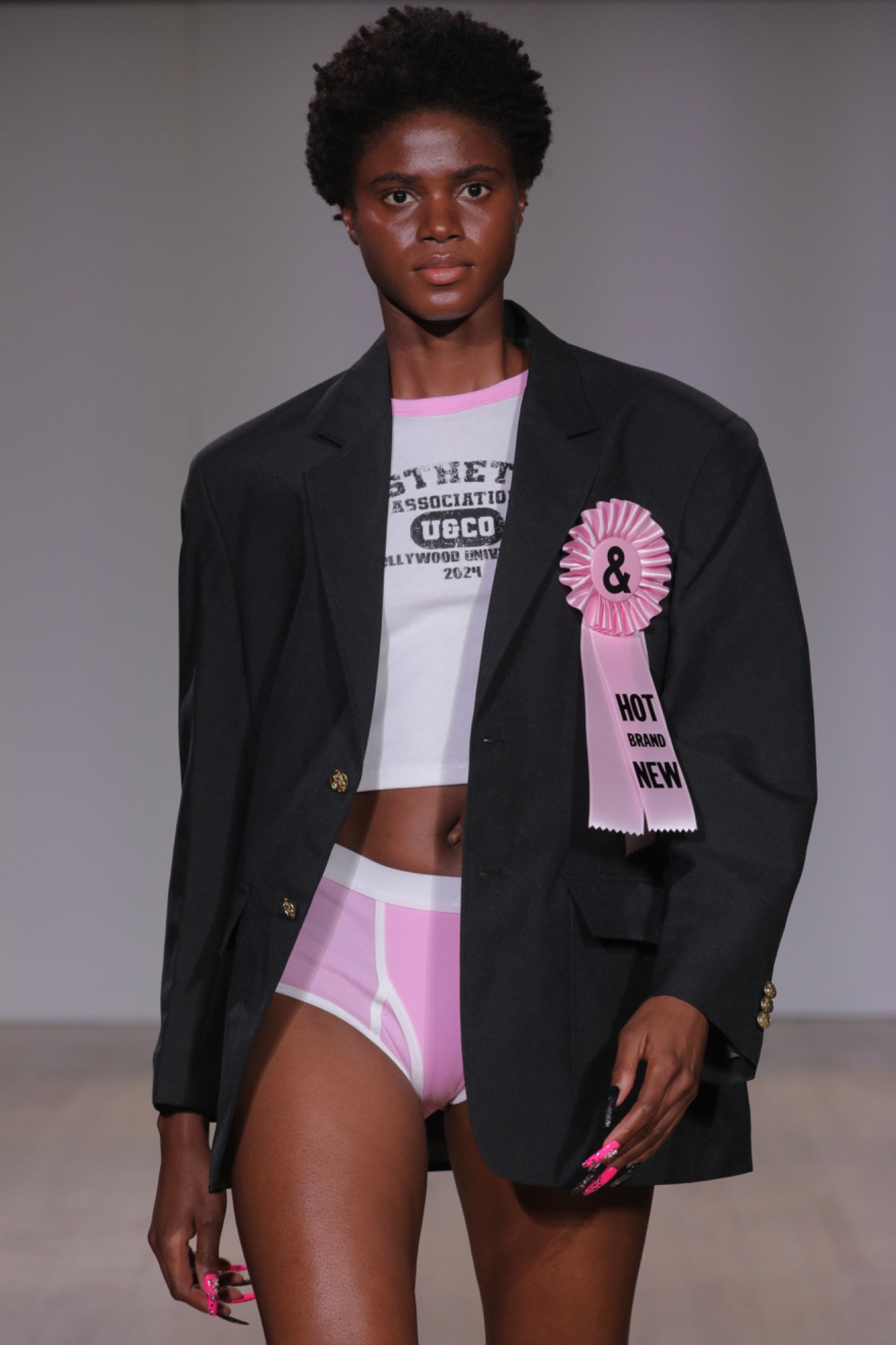 A model walking the runway wearing a white top, pink shorts, black blazer and pink embellishment.