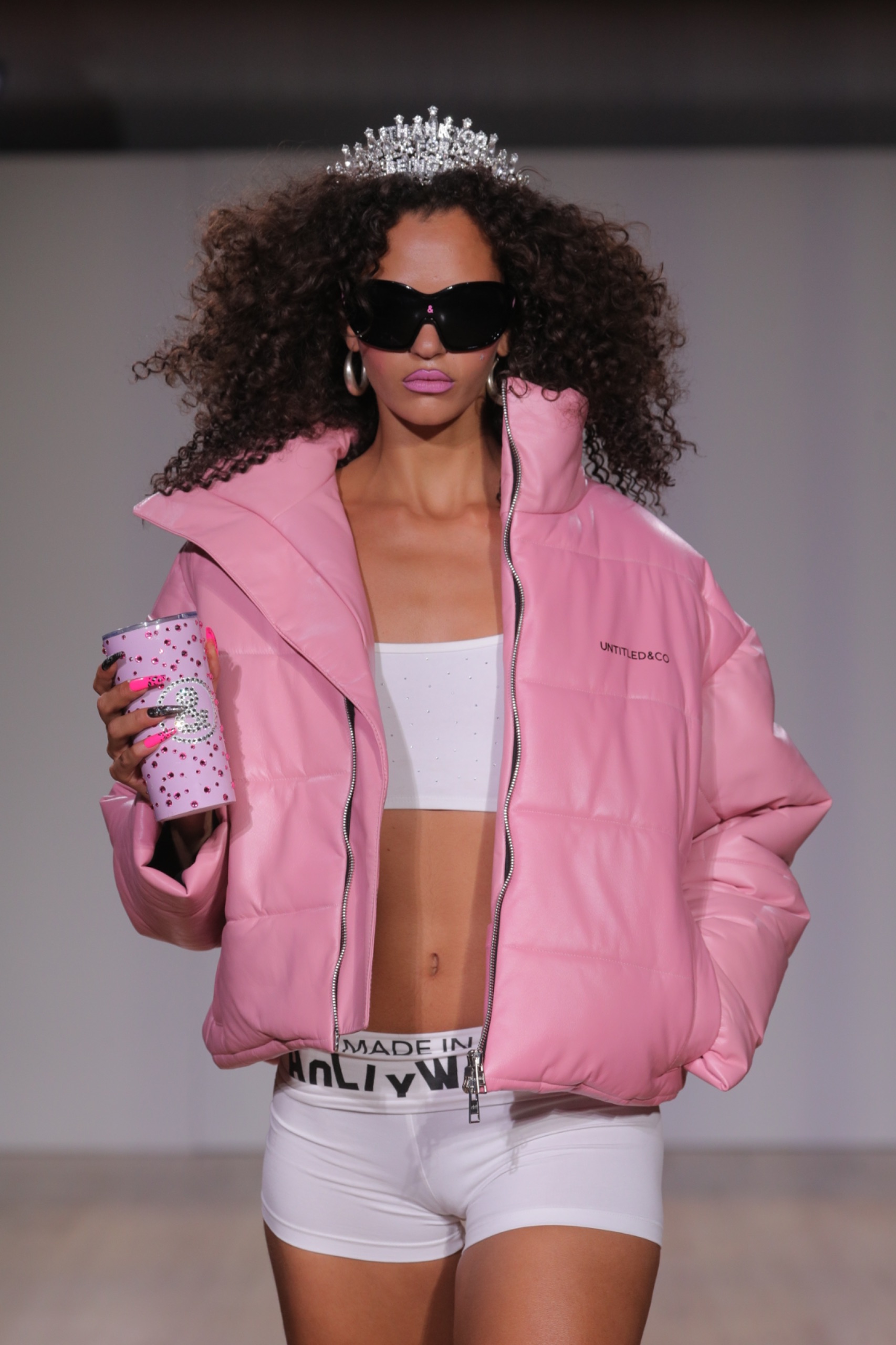 A model walking the runway wearing a tiara, pink coat, white top and is holding a drink cup.