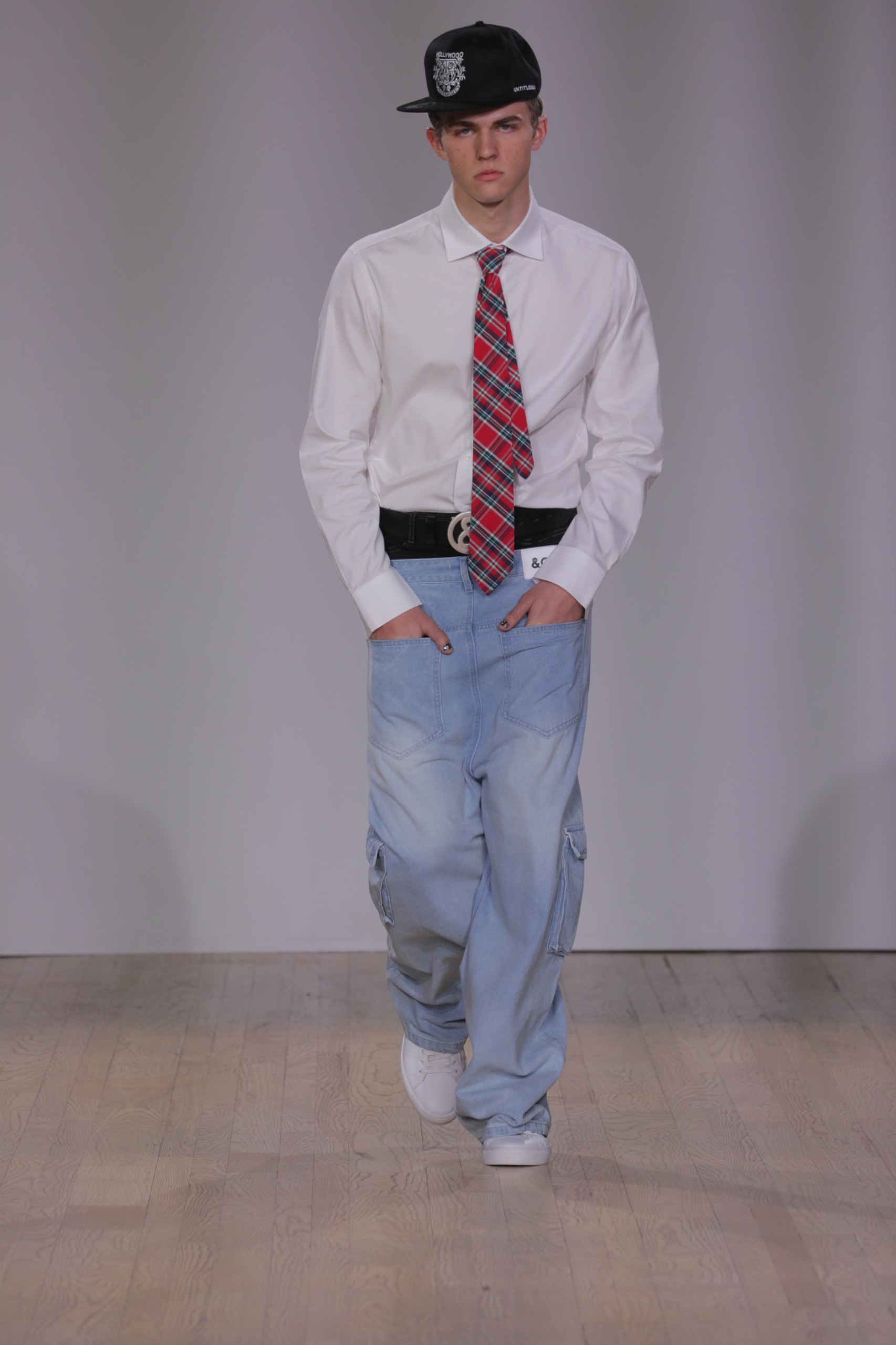 A model walking the runway wearing a black cap, white shirt, tie and blue jeans.