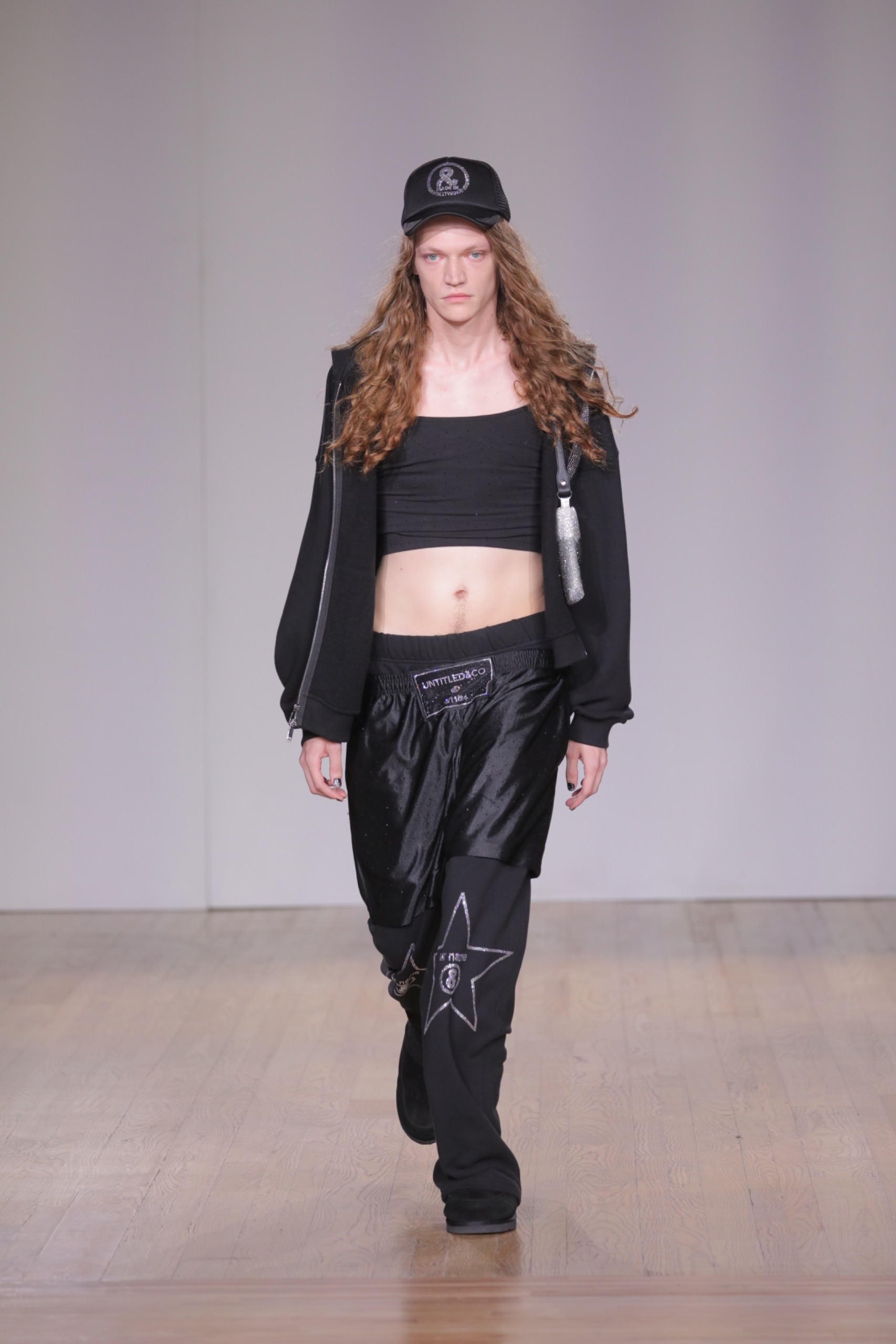 A model walking the runway wearing a black top, cap, jacket and pants.