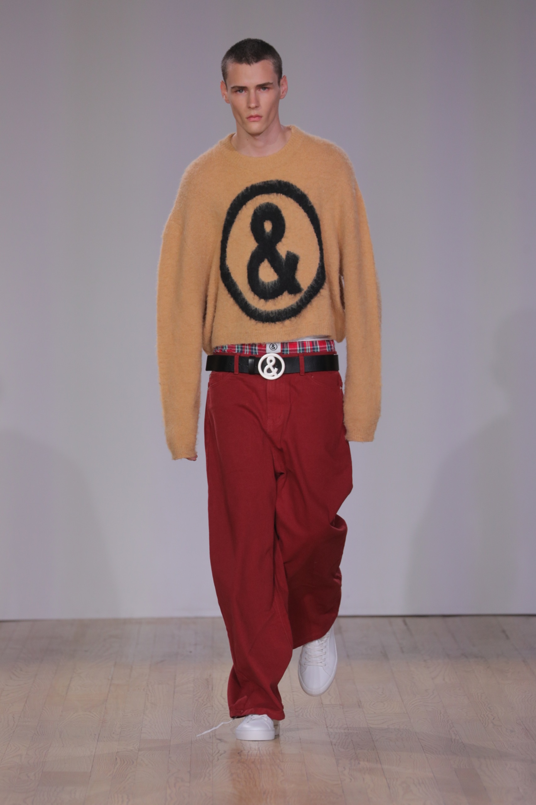 A model walking the runway wearing an orange jumper with a logo and red pants.