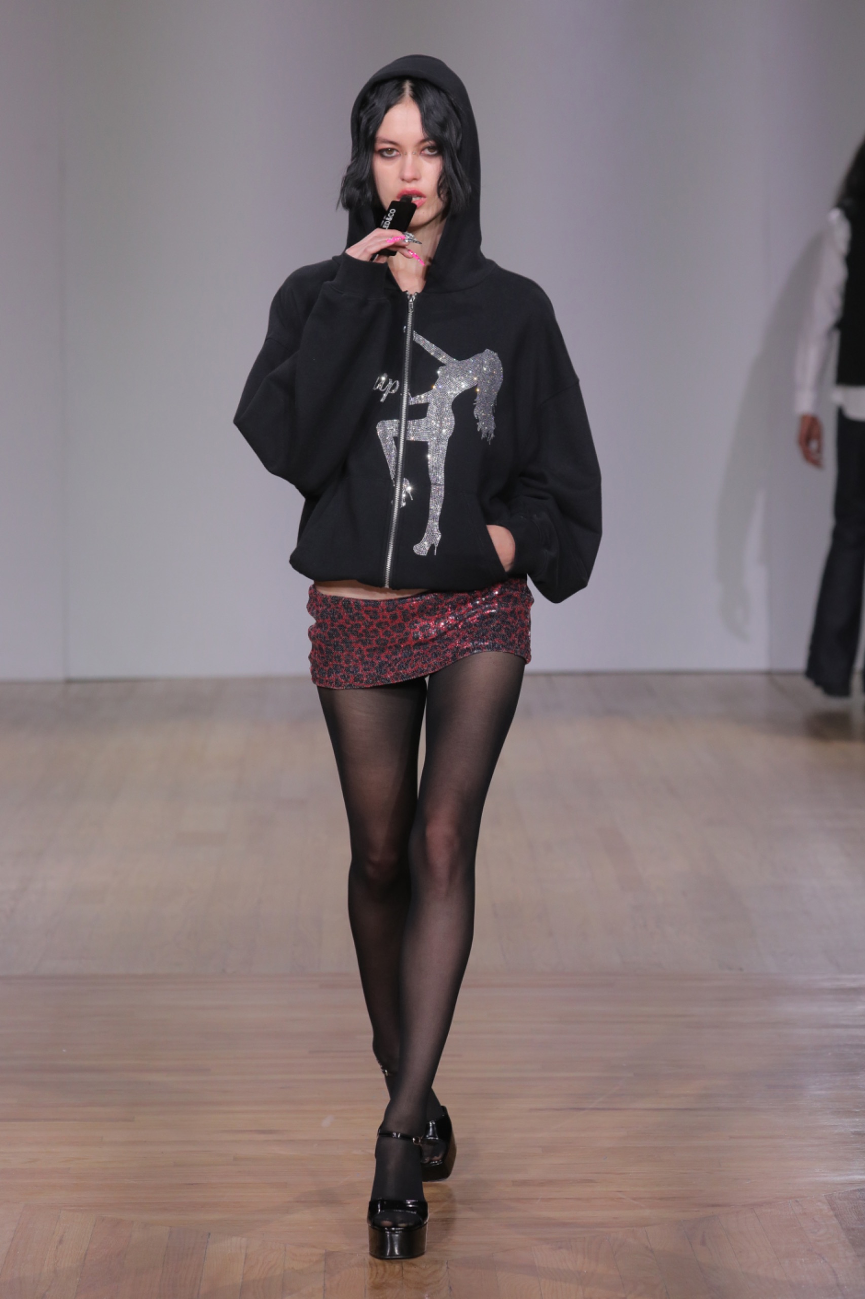 A model walking the runway wearing a black jacket with white detailing, pink skirt and black tights.