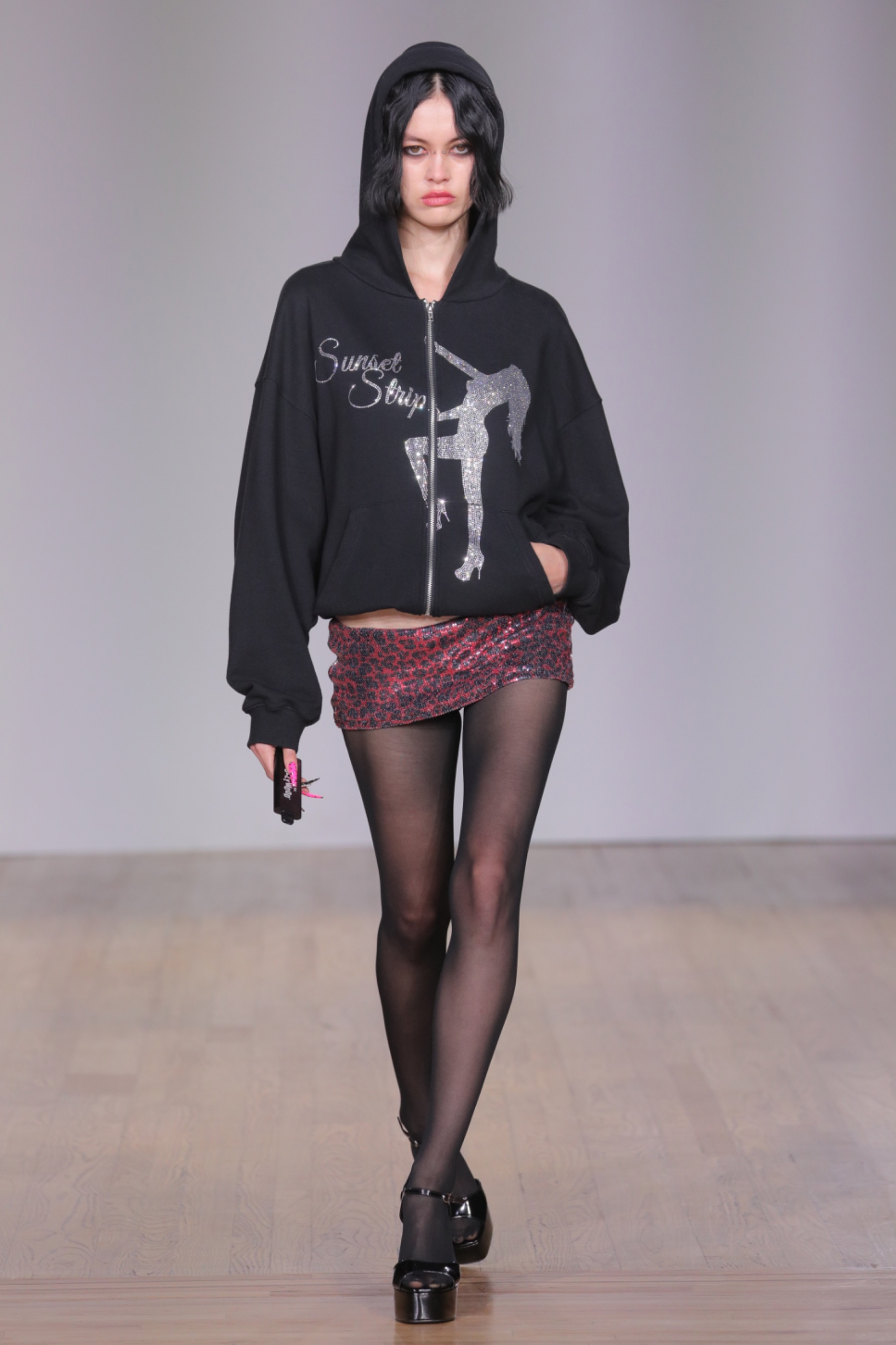 A model walking the runway wearing a black jacket with white detailing, pink skirt and black tights.