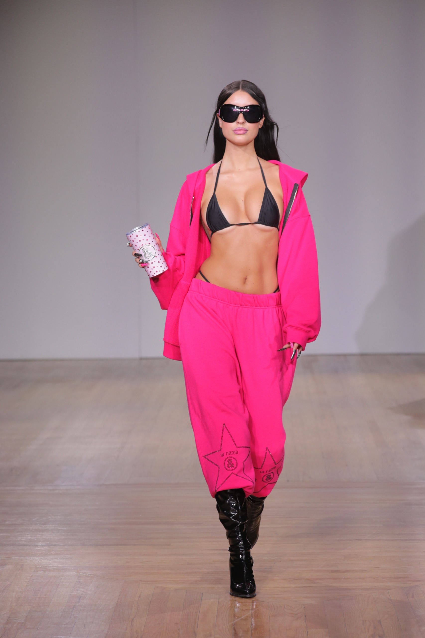 A model walking the runway wearing black sunglasses, a black bikini top, a pink jacket and pink pants.