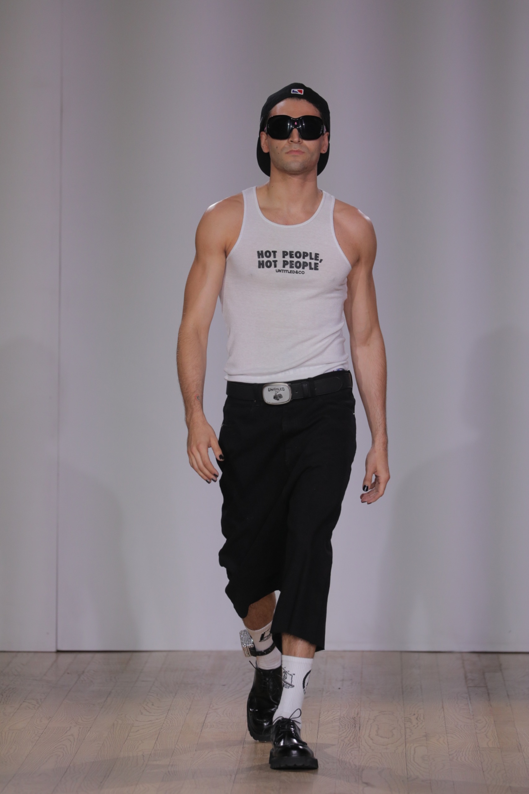 A model walking the runway wearing black sunglasses, a white tank with black lettering and black pants.