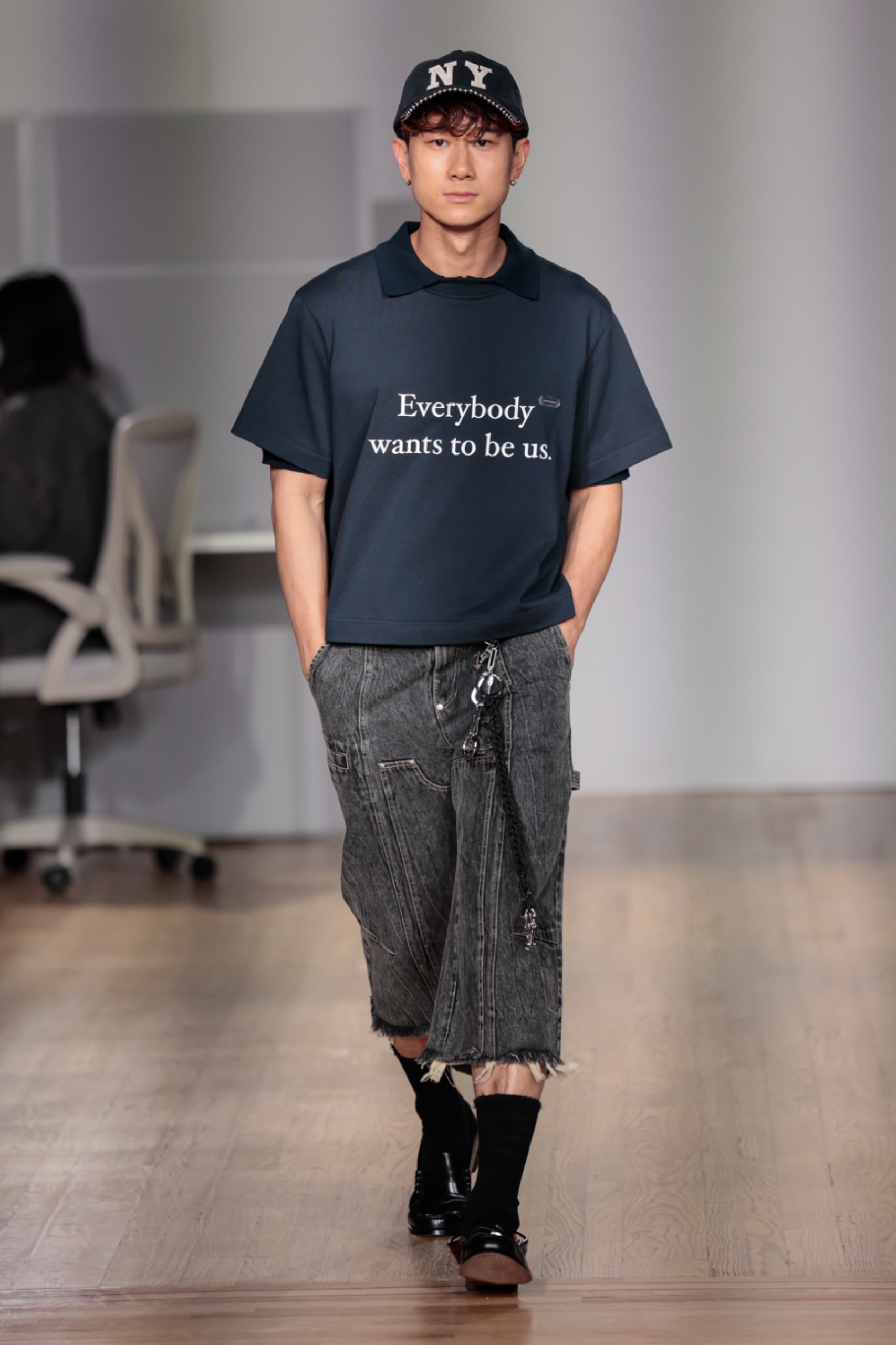 A model walking the runway wearing a t-shirt with white writing and dark grey pants.