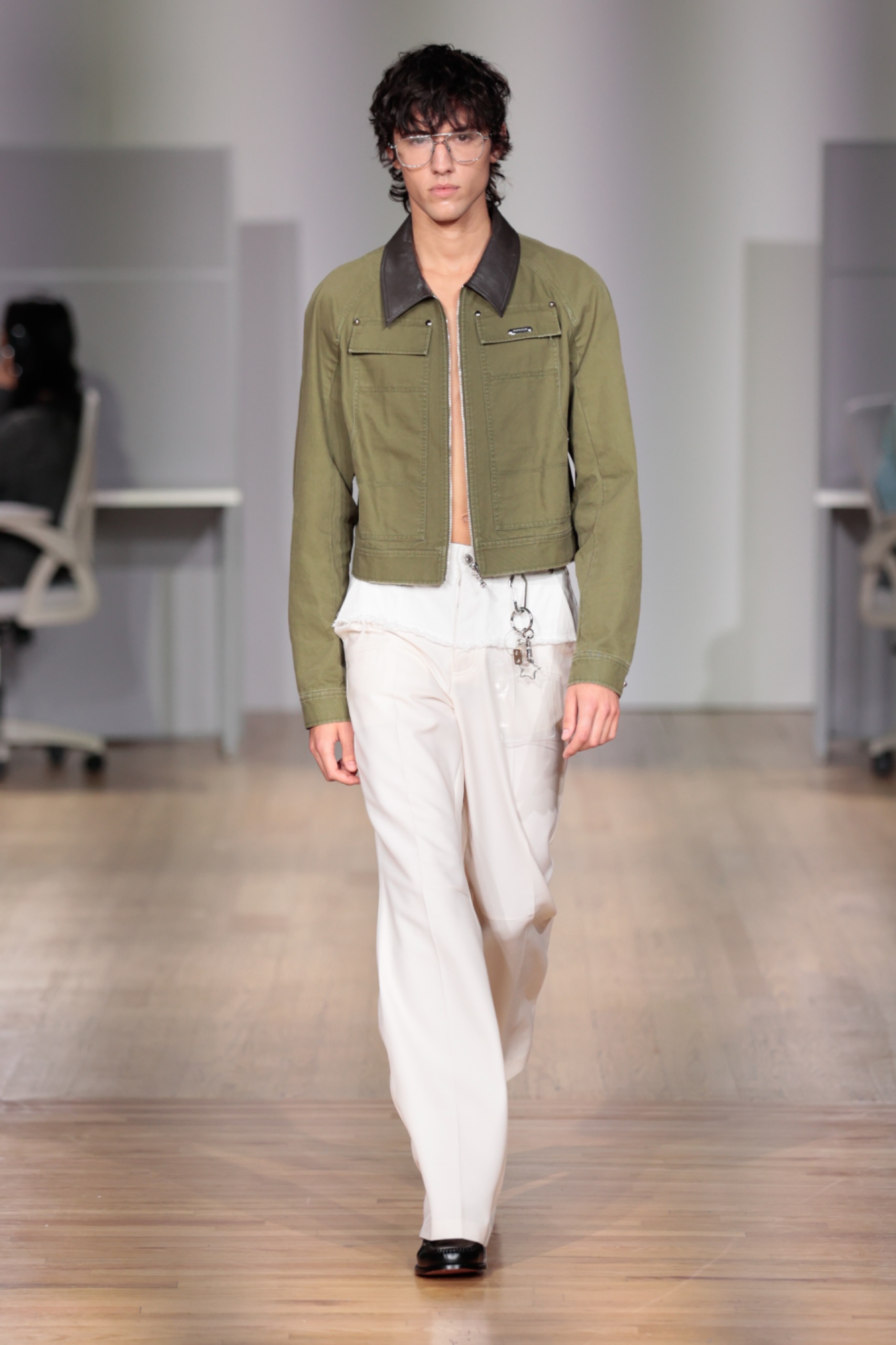 A model walking the runway wearing a green jacket and white pants.