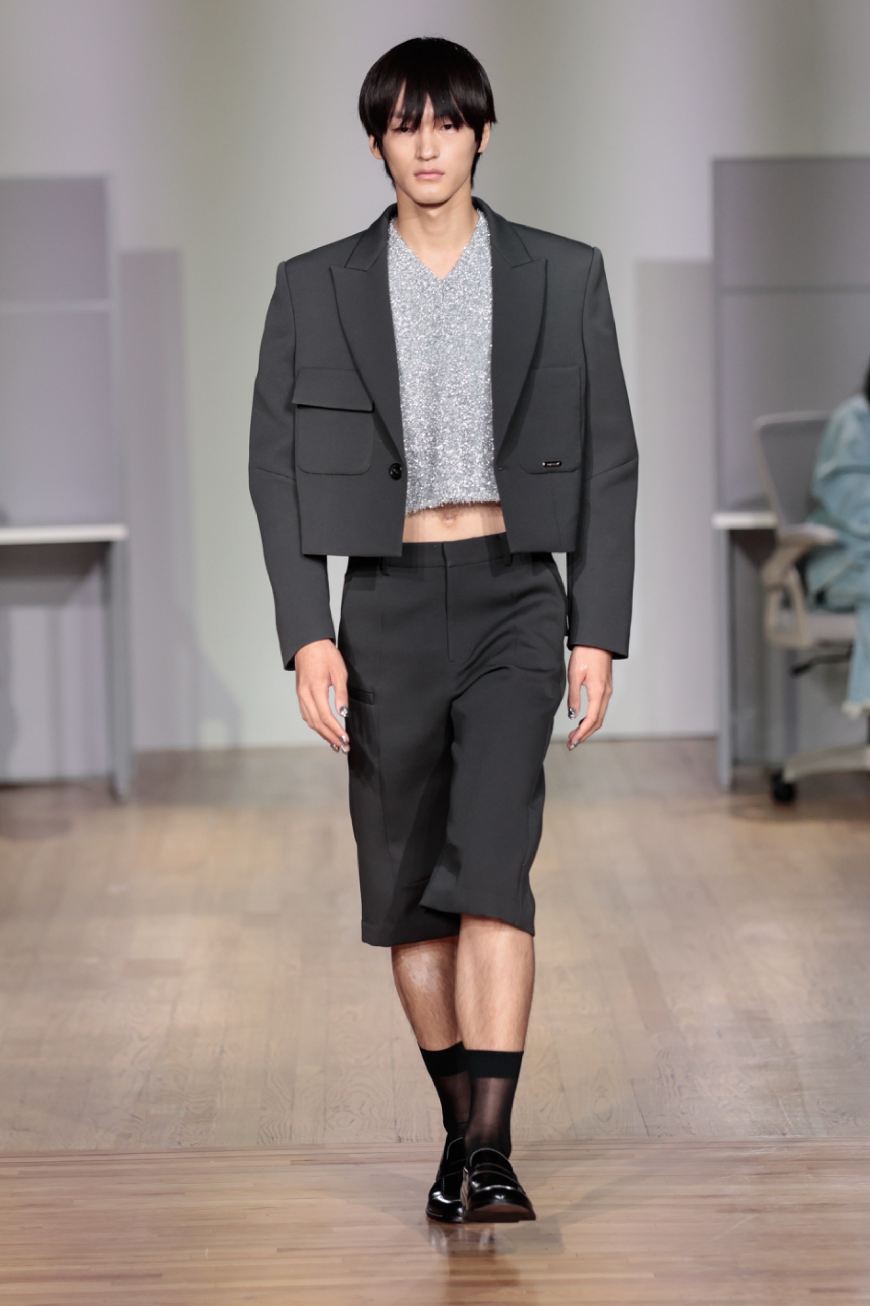 A model walking the runway wearing a black blazer, grey top and black shorts.