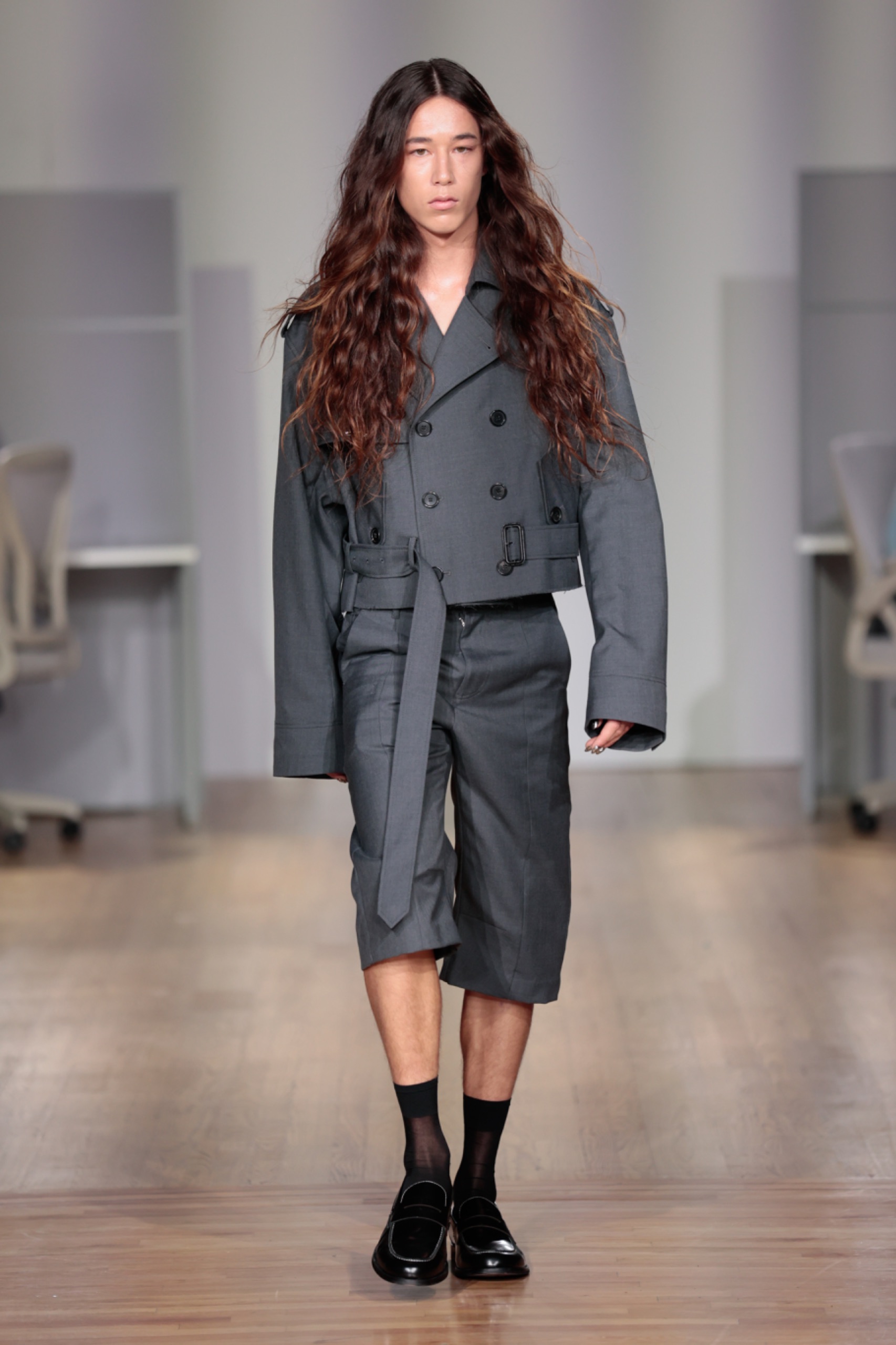 A model walking the runway wearing a grey jacket and shorts.