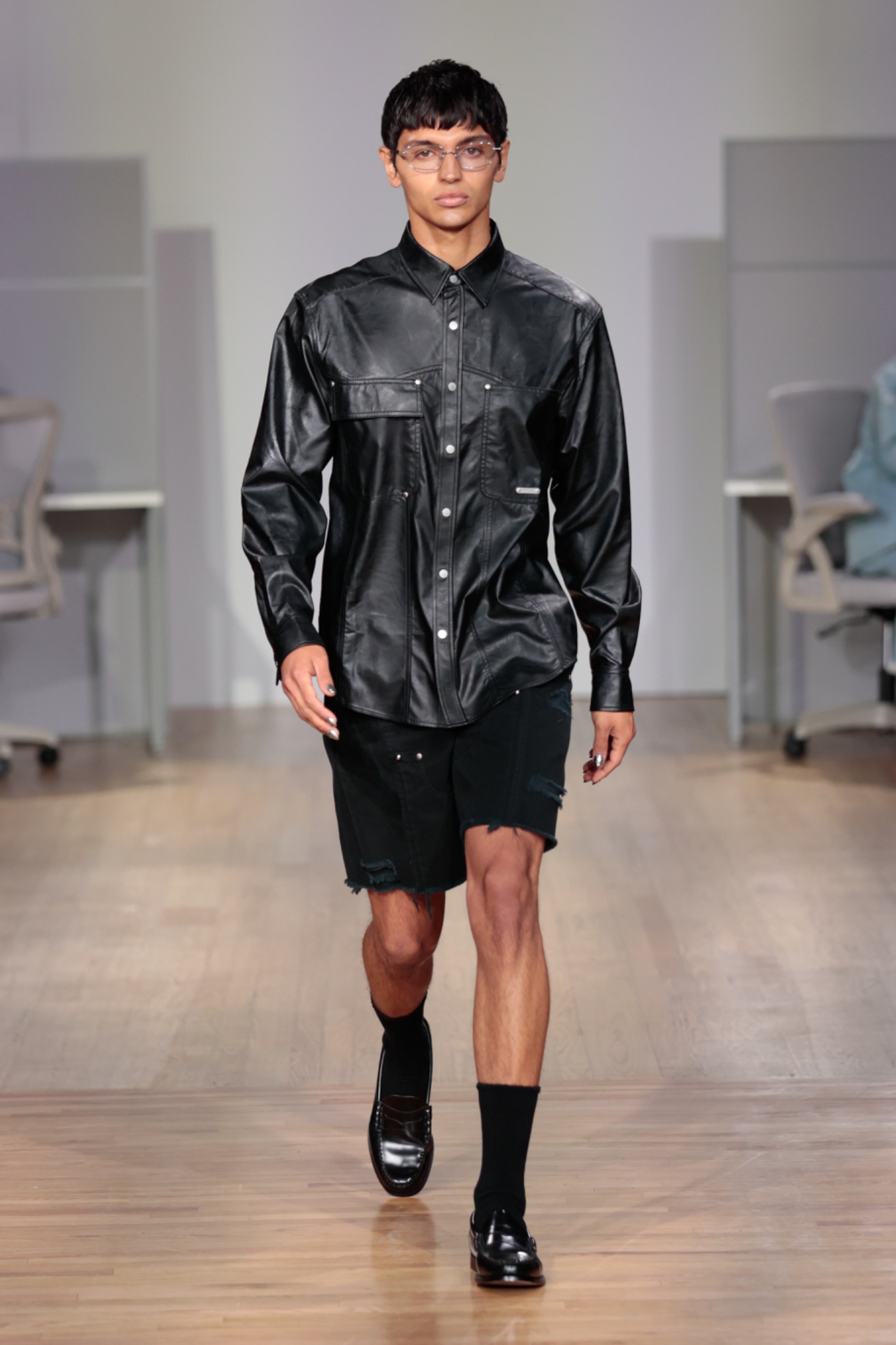 A model walking the runway wearing a black jacket, black shorts and black shoes.