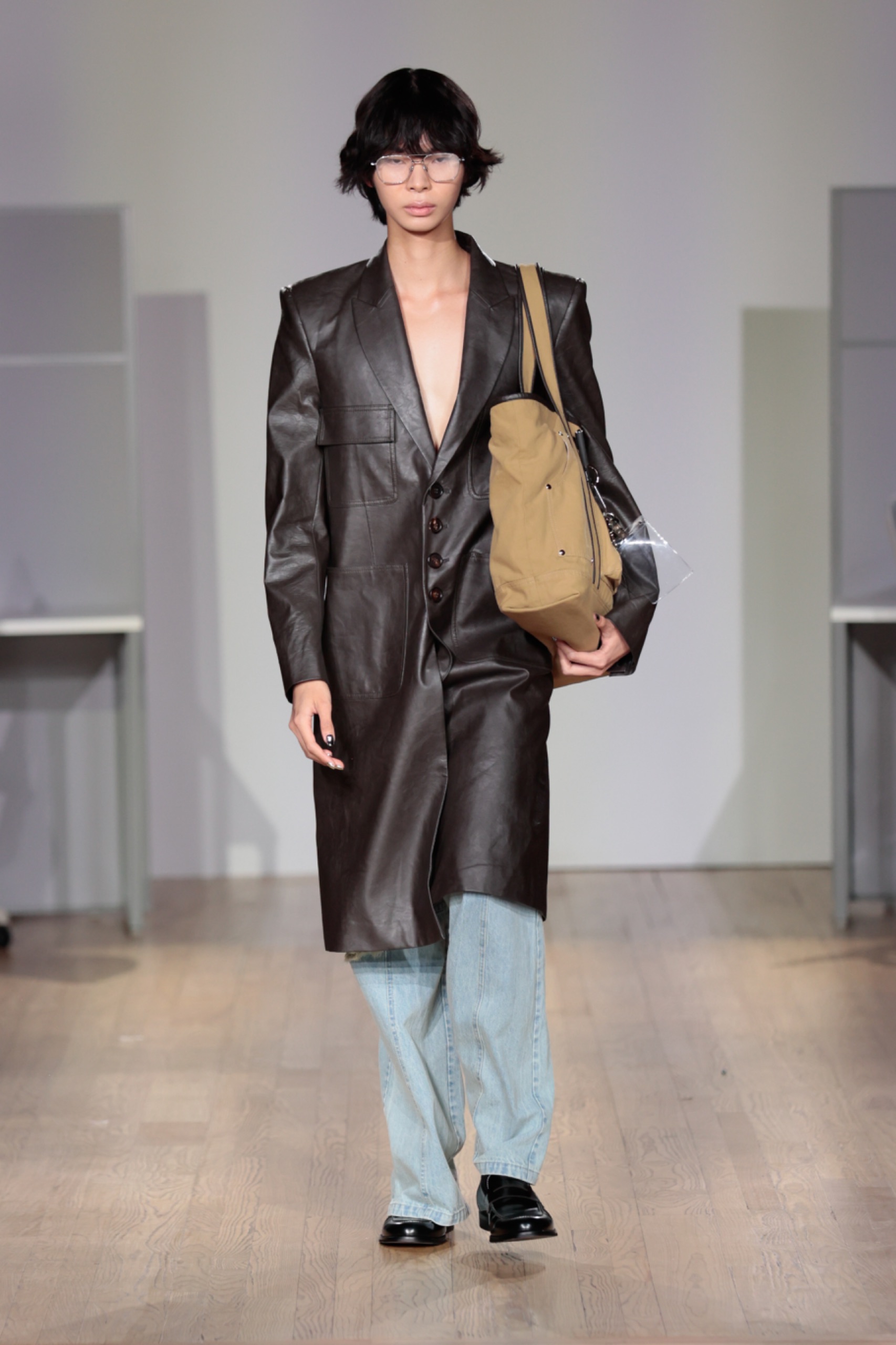 A model walking the runway wearing a long black coat, bag and light blue jeans.