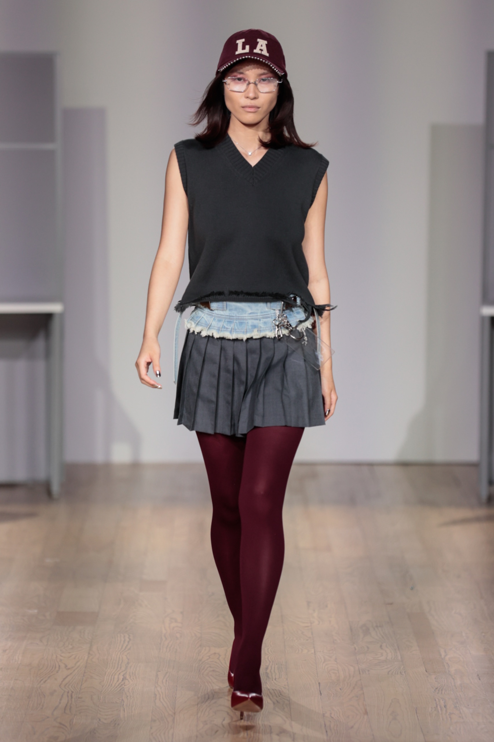 A model walking the runway wearing a black vest, grey skirt and maroon tights.