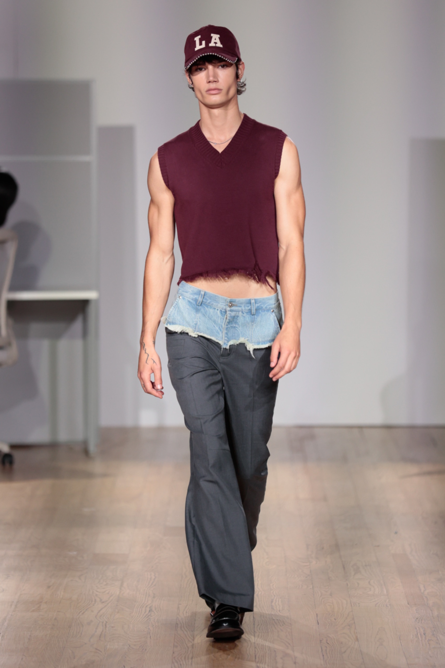 A model walking the runway wearing a maroon vest and grey pants with blue denim detail.