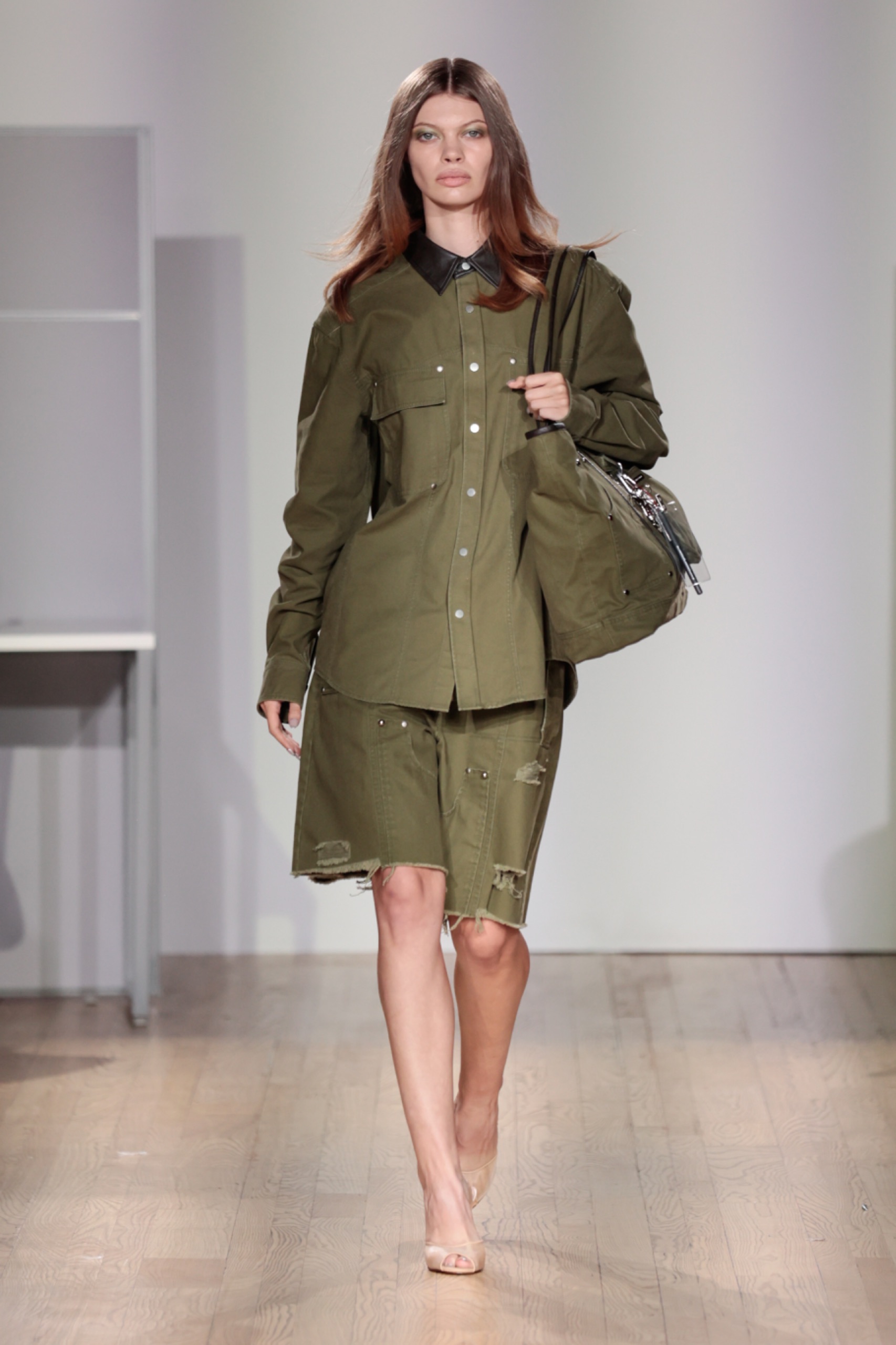 A model walking the runway wearing a green jacket, skirt and bag.