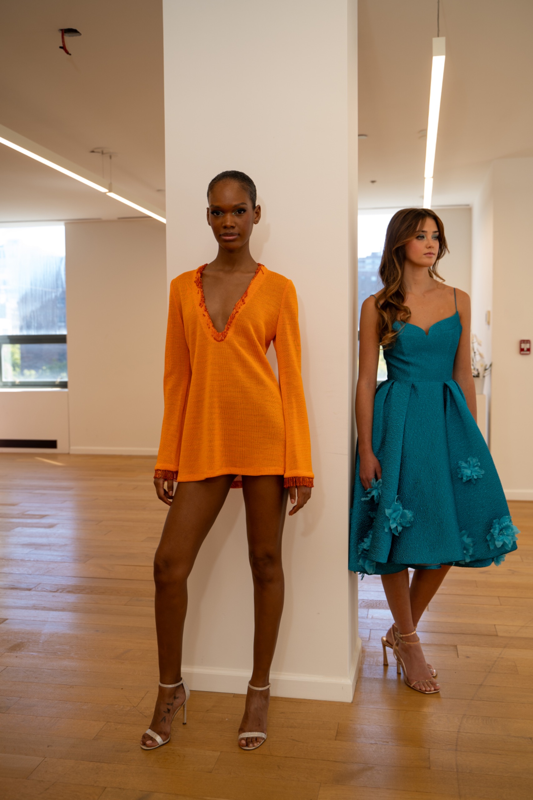 2 models. The model on the left is wearing an orange dress. The model on the right is wearing a long green/blue dress.