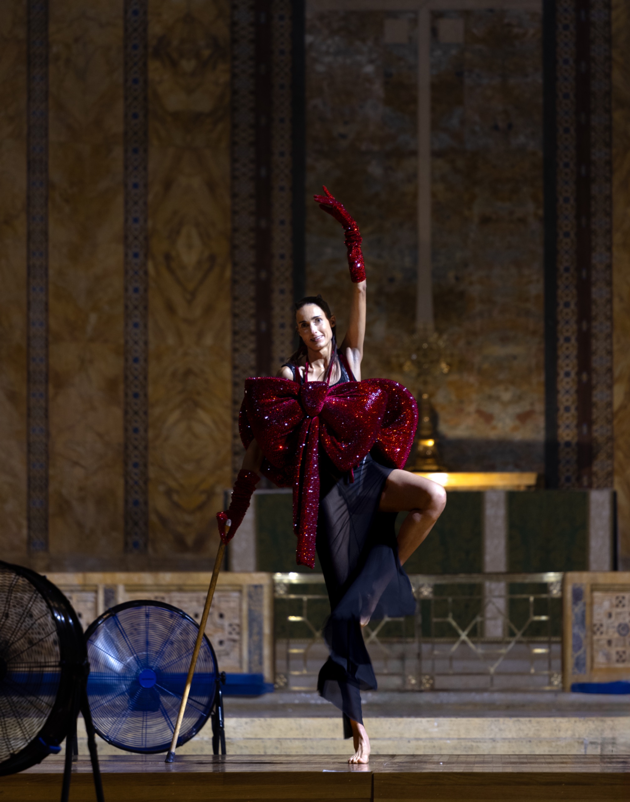 Model performs ballet while wearing long black dress with red bow.