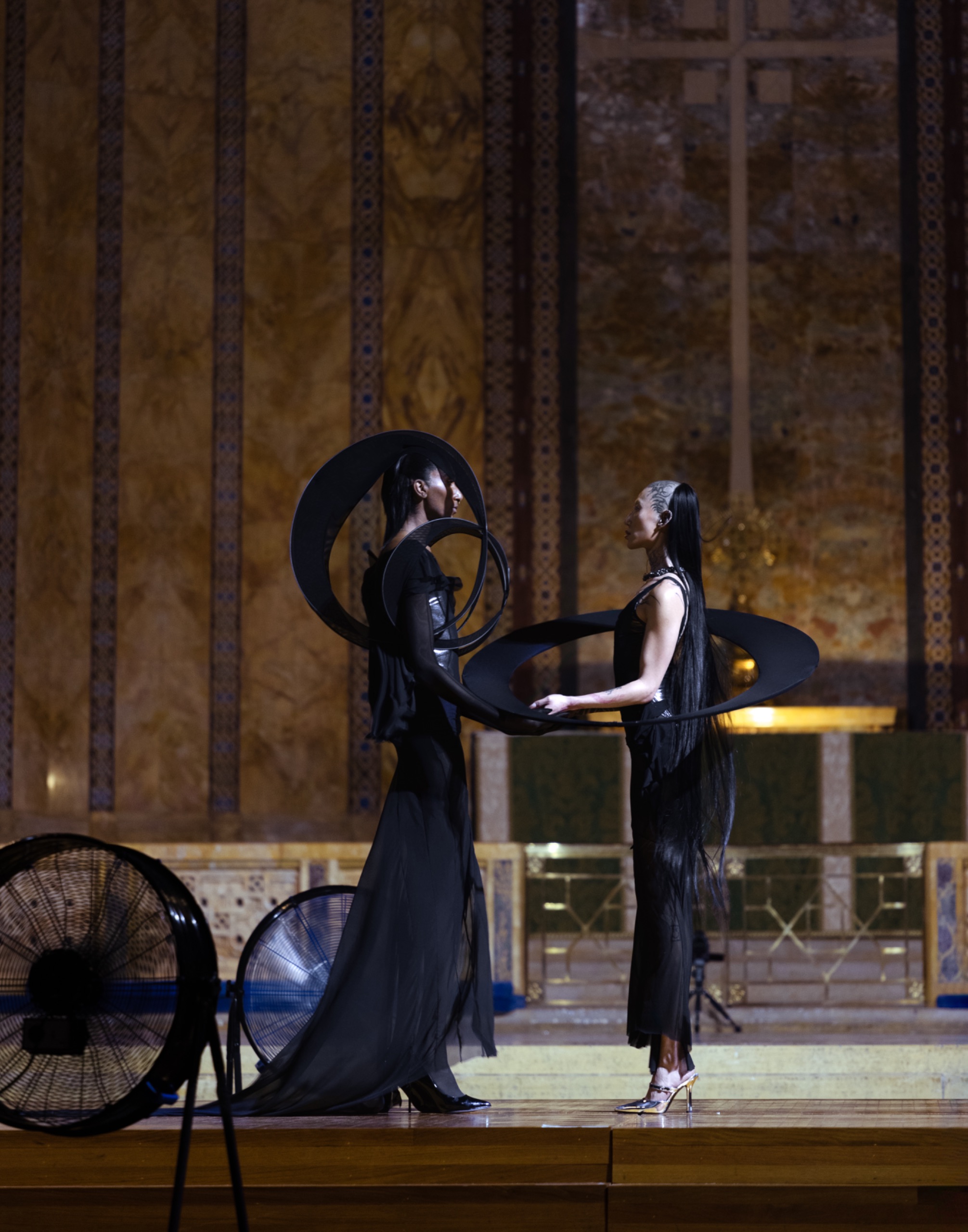 Two models hold each other’s arms, posing with long black dresses on.