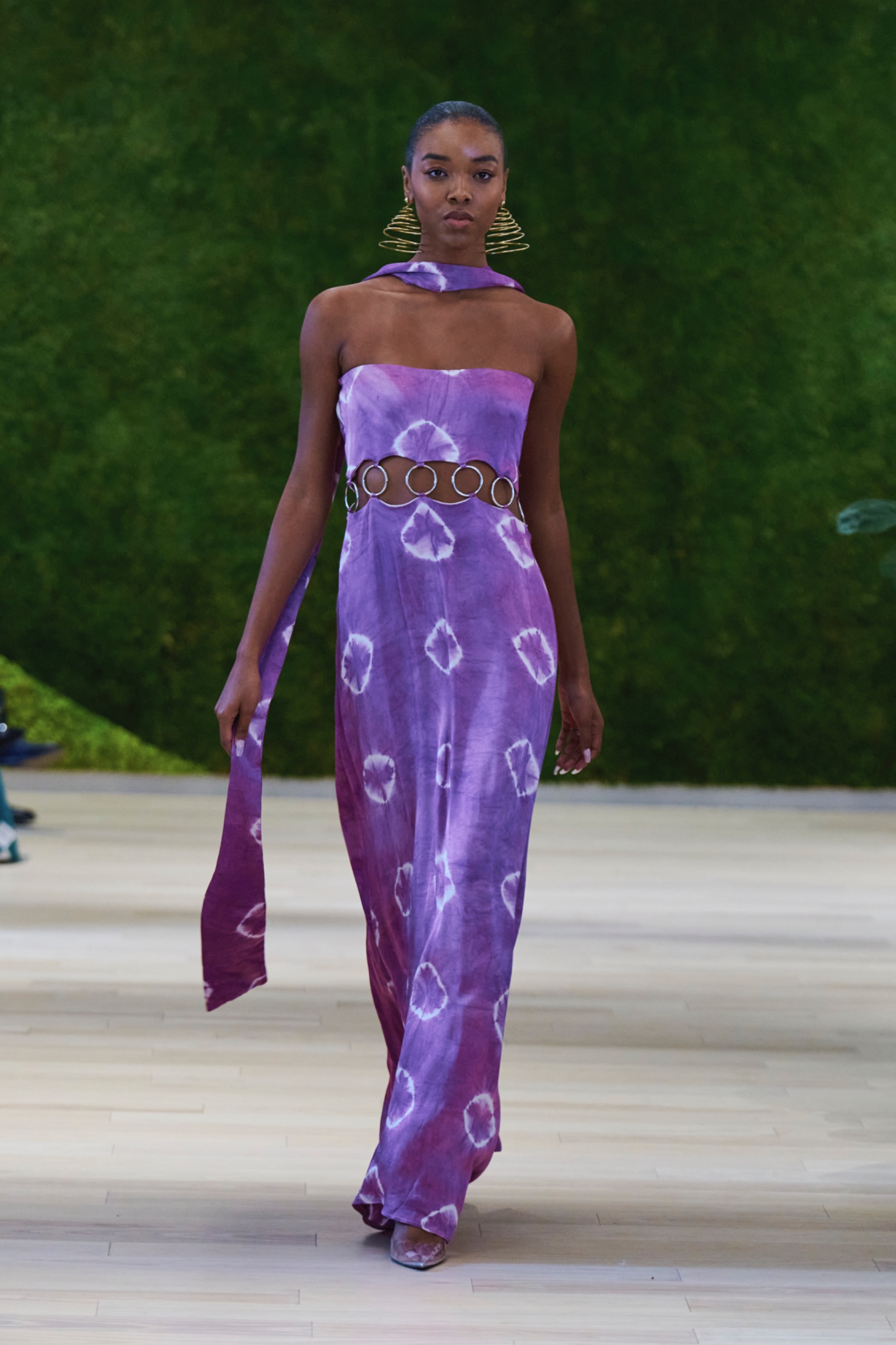 A model walking the runway wearing a purple top and skirt with swirled pattern.