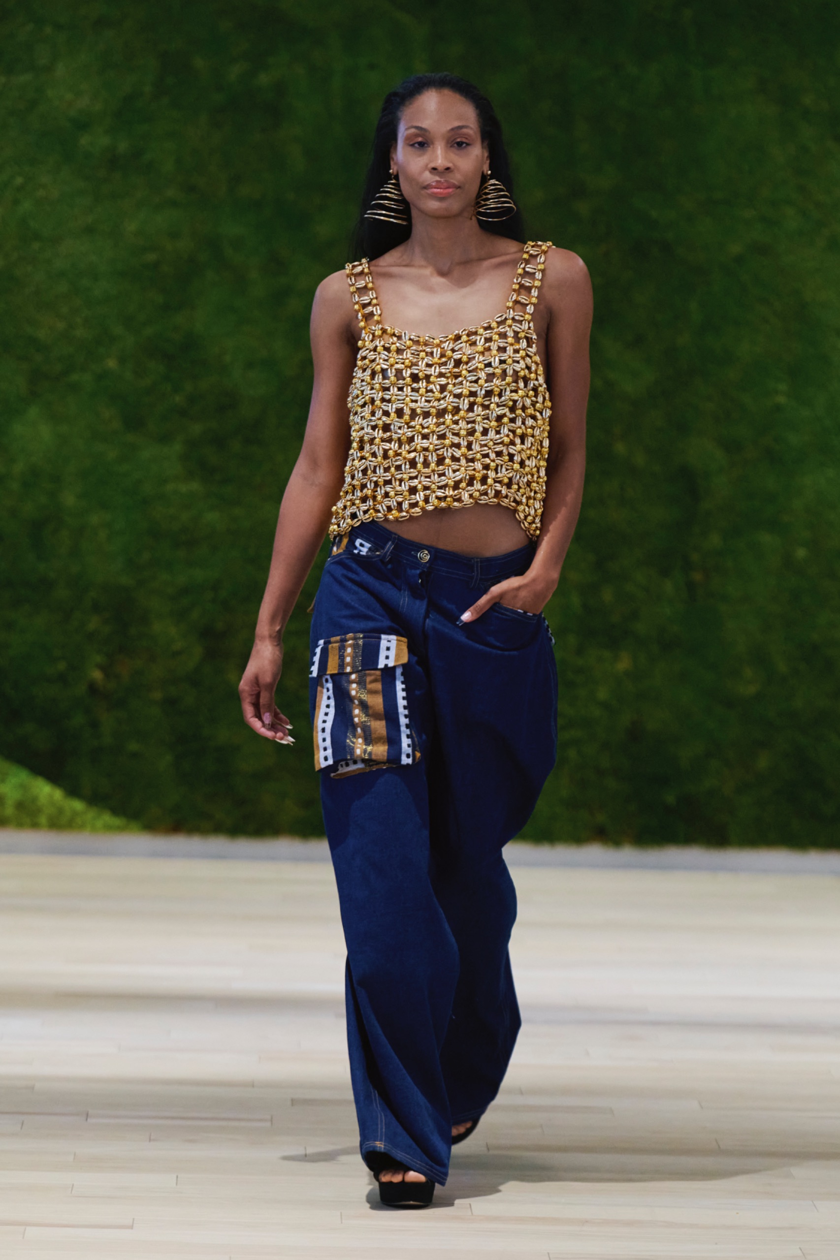 A model walking the runway wearing a gold top and blue pants with striped pocket.