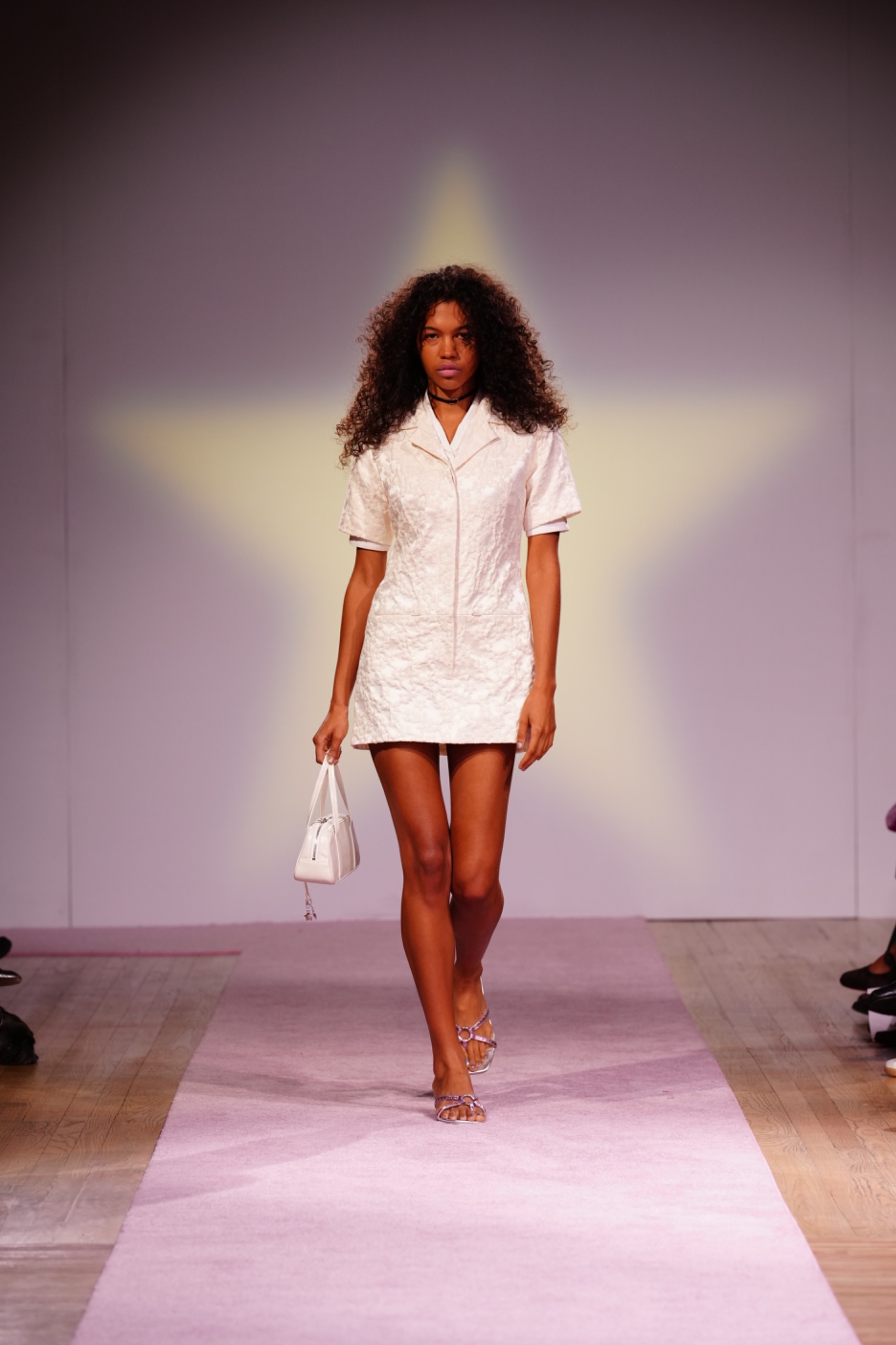 A model walking the runway wearing a white t-shirt dress with a white bag.