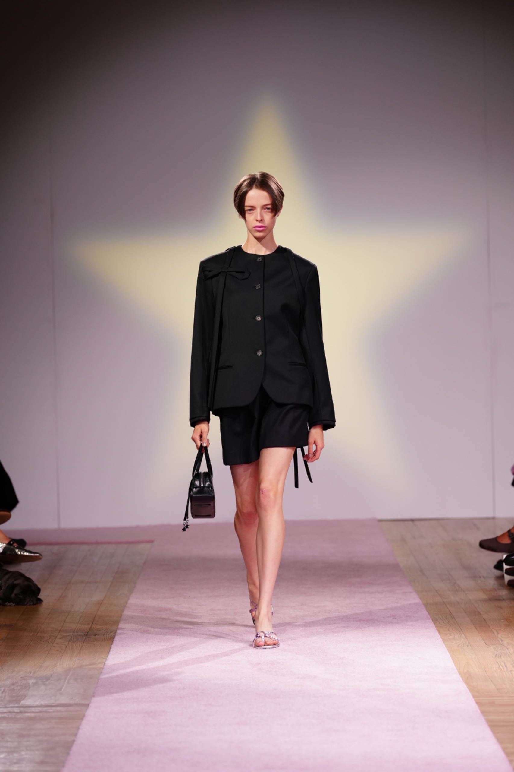 A model walking the runway wearing a black blazer, black skirt and black bag.