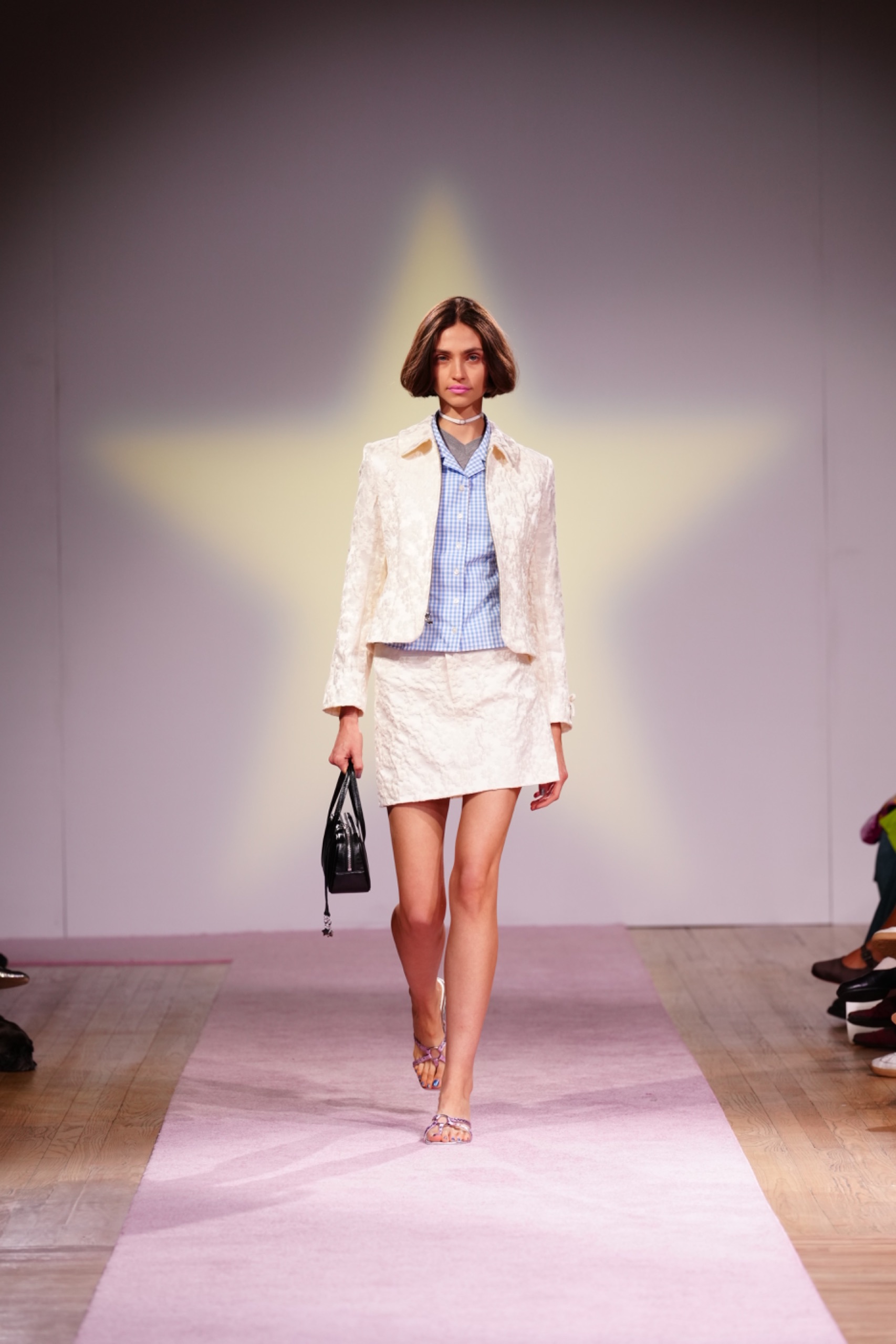 A model walking the runway wearing a white blazer, skirt and blue shirt.