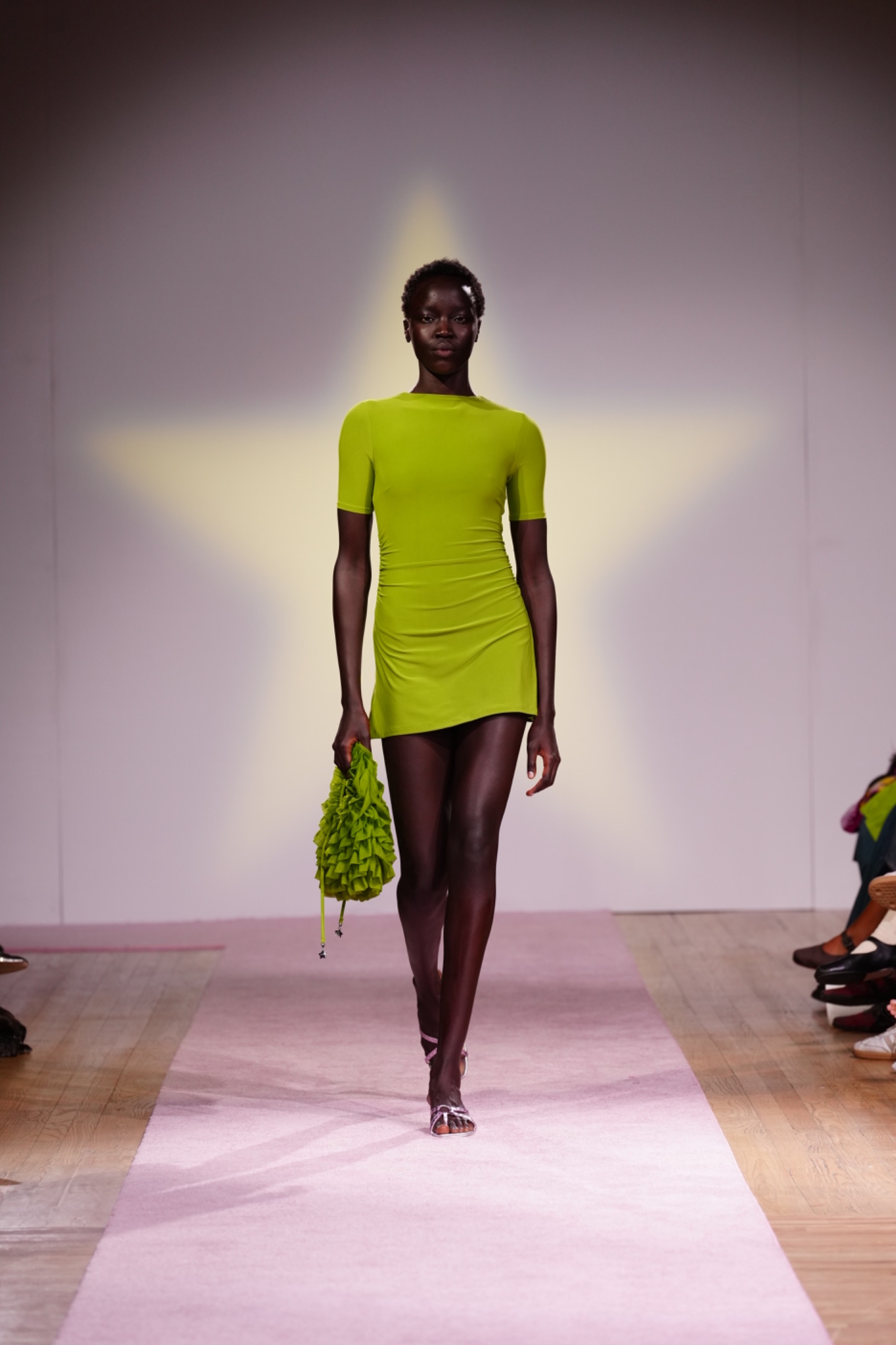 A model walking the runway wearing a green dress and bag.