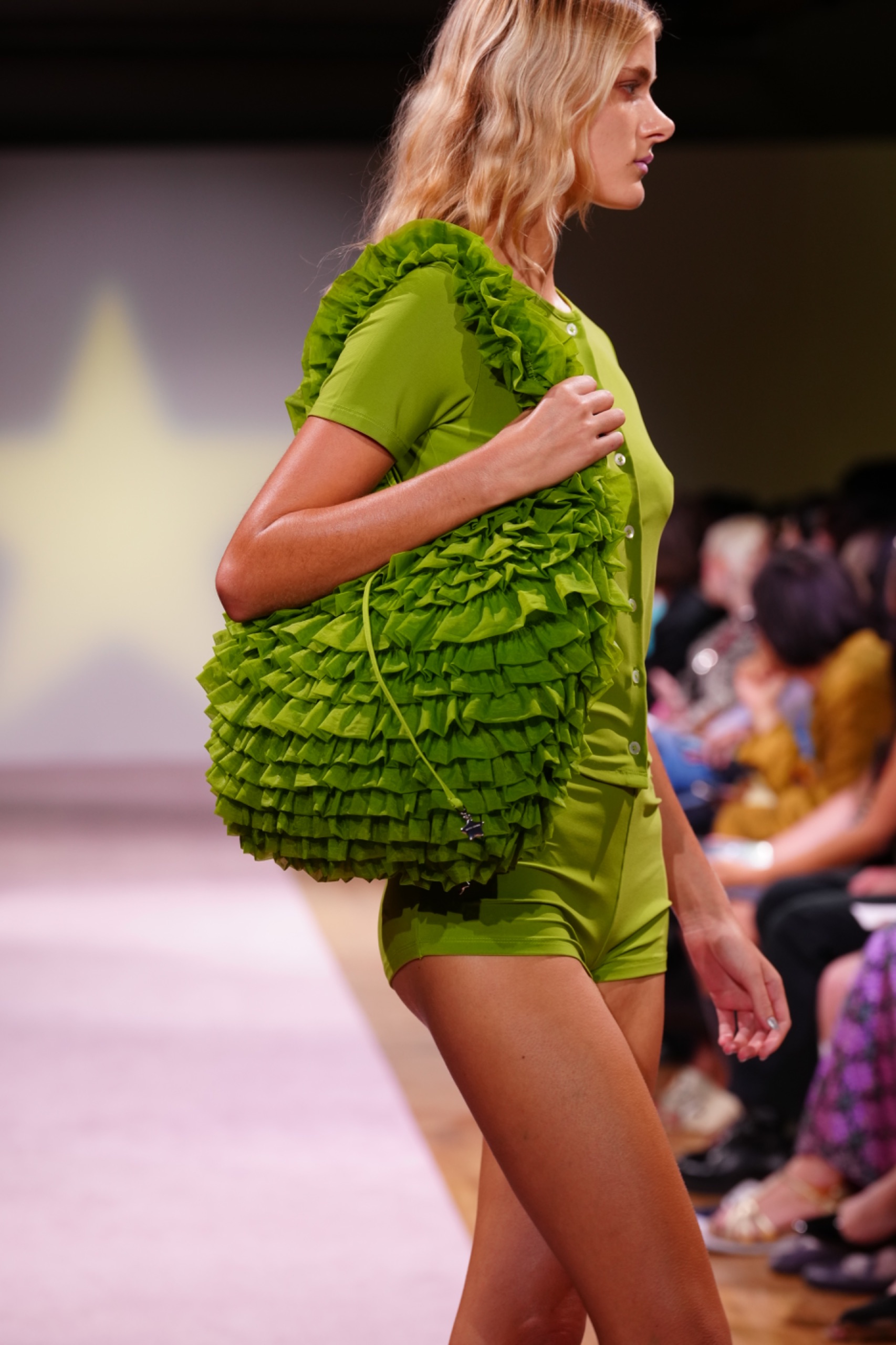 A model walking the runway wearing a green cardigan and shorts with a green bag.