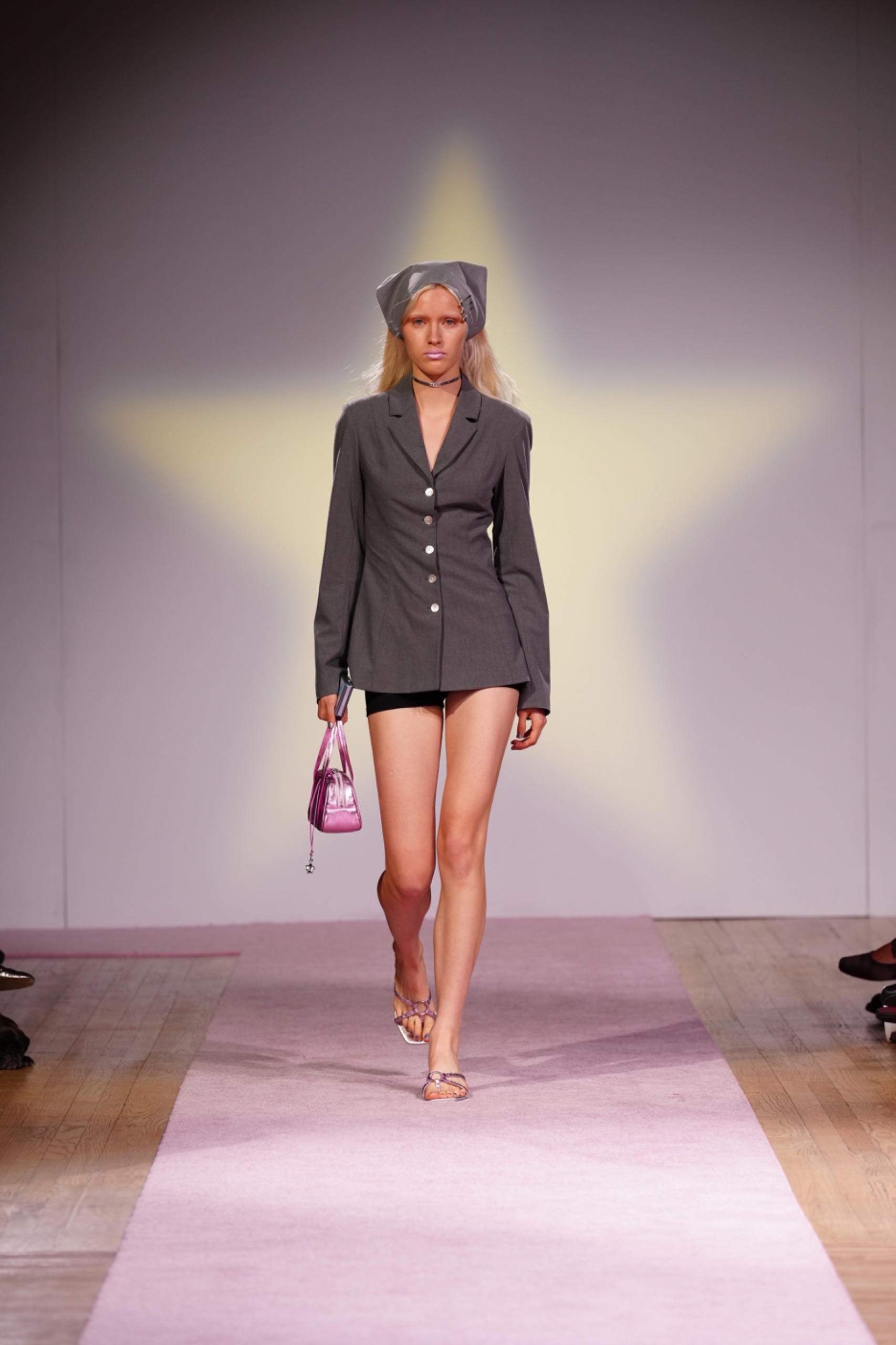 A model walking the runway wearing a grey blazer with a pink bag.