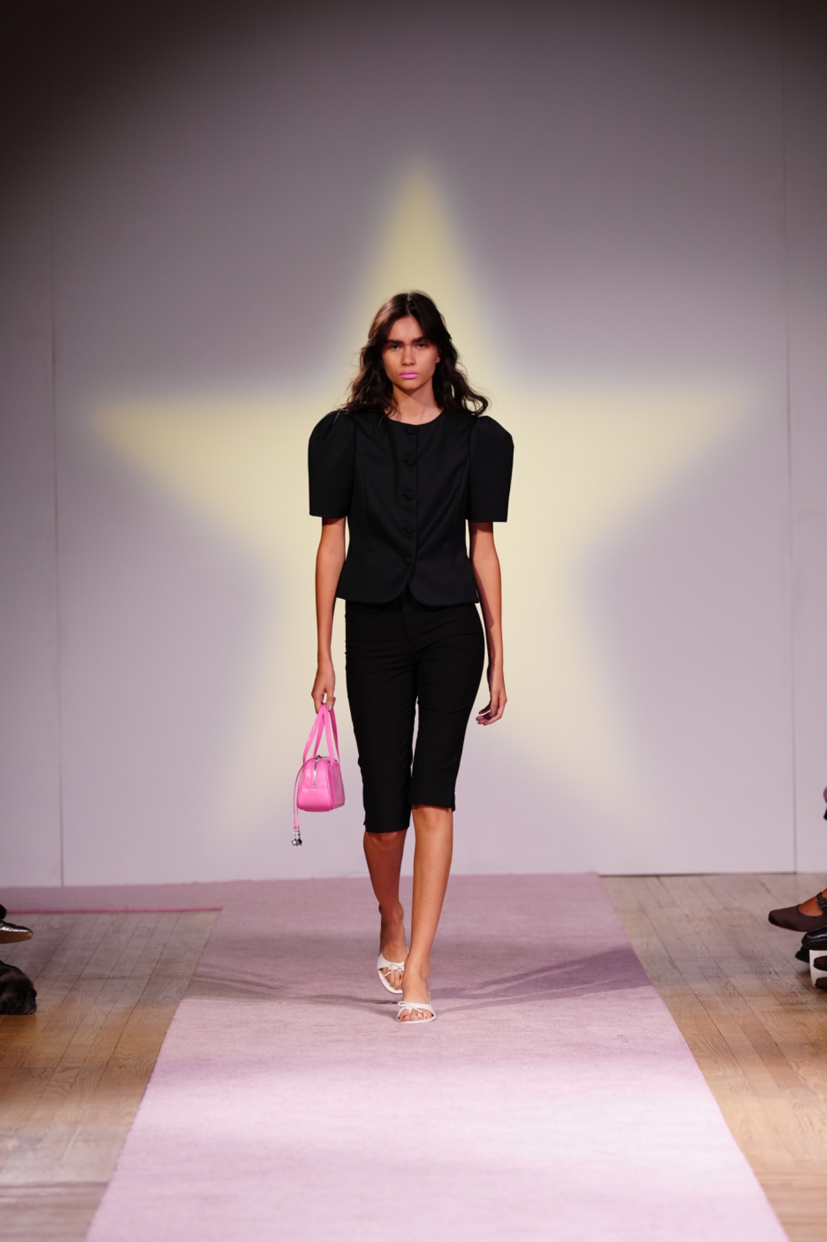 A model walking the runway wearing a black top and black pants with a pink bag.