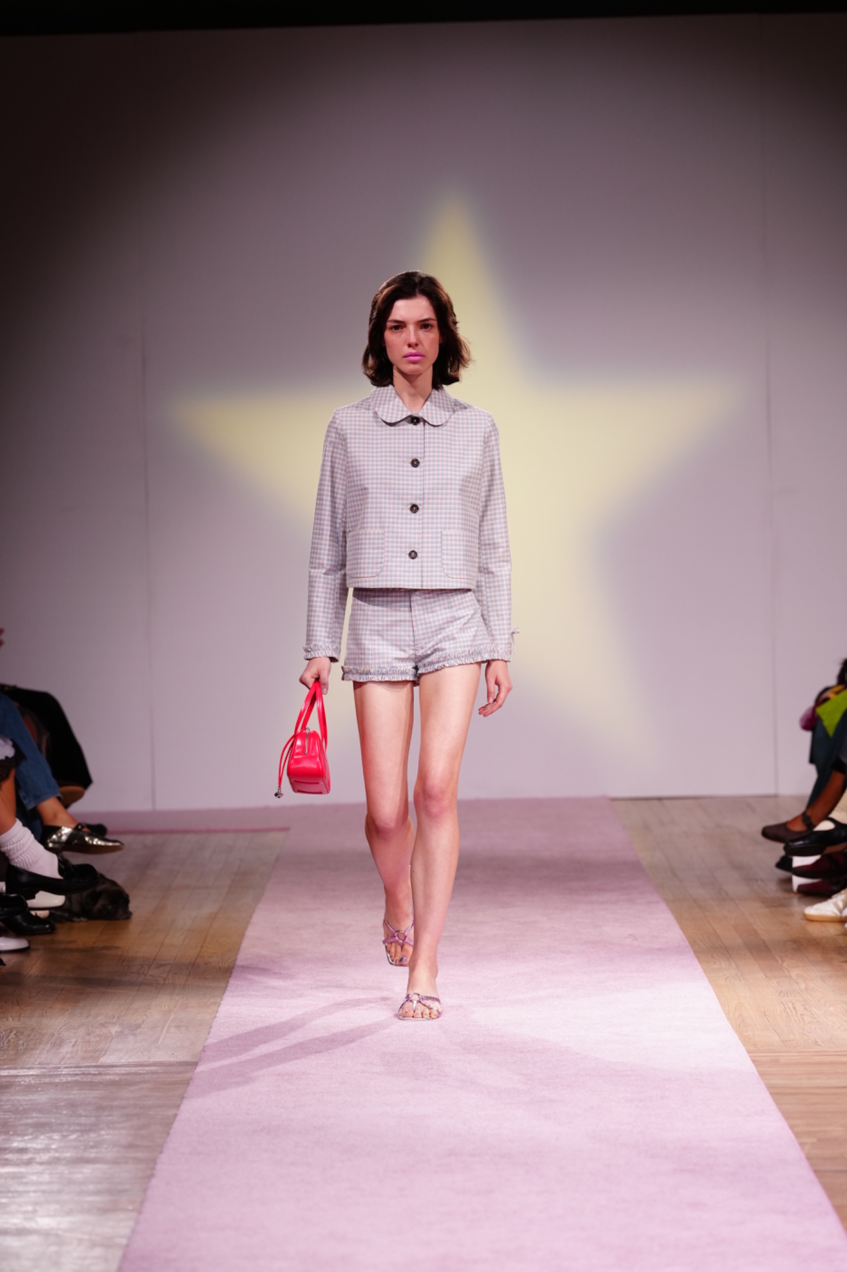 A model walking the runway wearing a grey blazer and shorts with a pink bag.