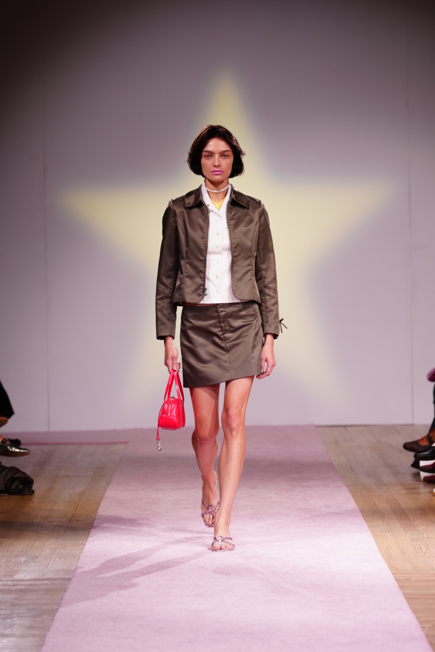 A model walking the runway wearing a brown jacket, skirt, white shirt and pink bag.