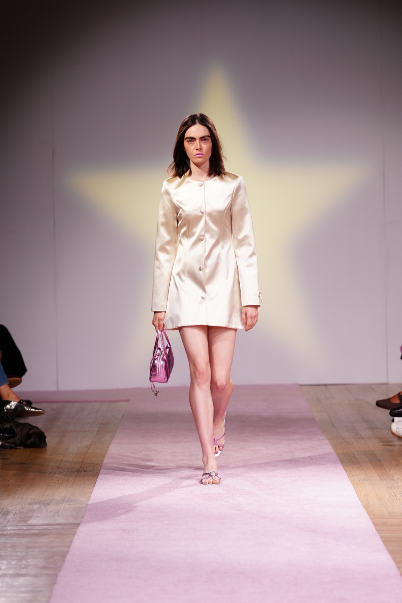 A model walking the runway wearing a ivory dress with a pink bag.